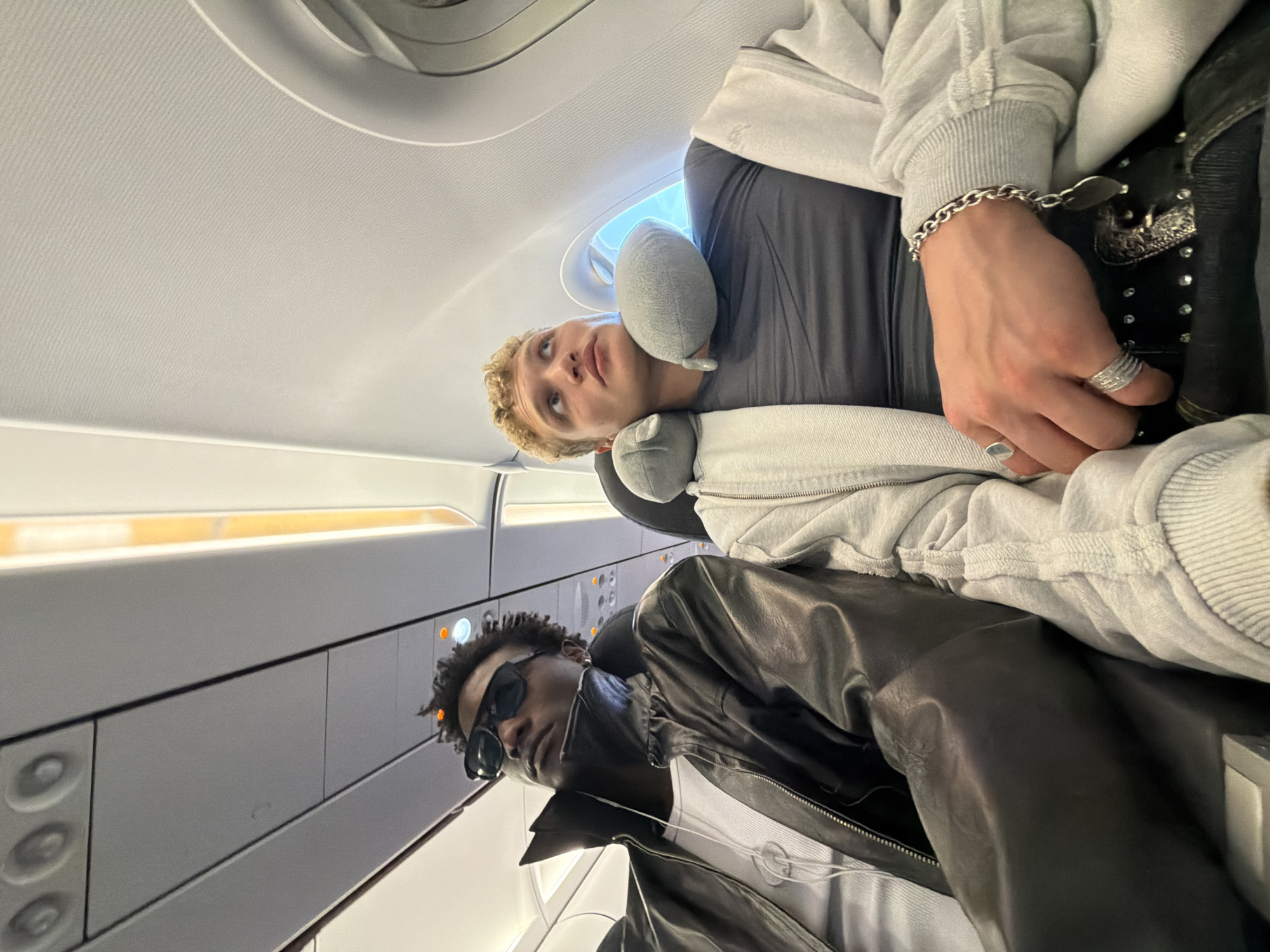 Two individuals are seated in an airplane cabin. The person on the left, wearing sunglasses and a black leather jacket over a white t-shirt, is looking upwards. The person on the right, with blonde curly hair and a grey t-shirt, is also looking upwards, with a grey neck pillow supporting their head. Both individuals appear to be passengers. The overhead compartment lights are illuminated, casting a warm glow on the white interior of the cabin. The image does not contain any specific elements that would allow for identification of the location as Crawley, United Kingdom.