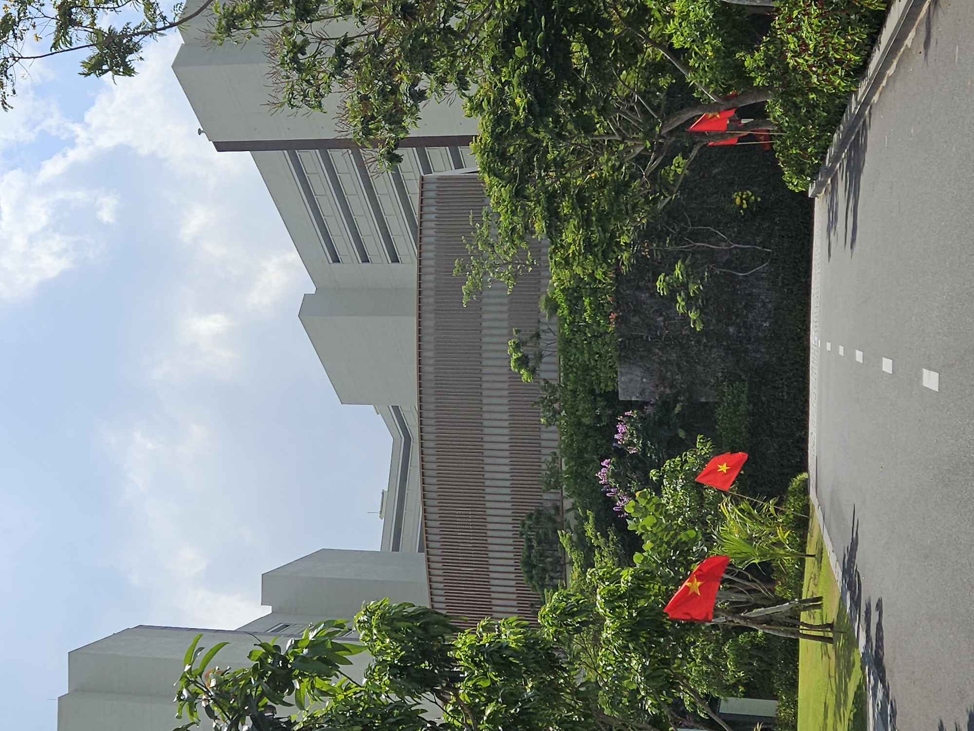 The image shows a building with a horizontally slatted facade next to a road with dotted white lines, with Vietnamese flags along the side of the road. The location is Bac Cam Ranh Ward, Vietnam. The building appears to be a multi-story structure, partially obscured by trees in the foreground. The sky is visible with some cloud cover.