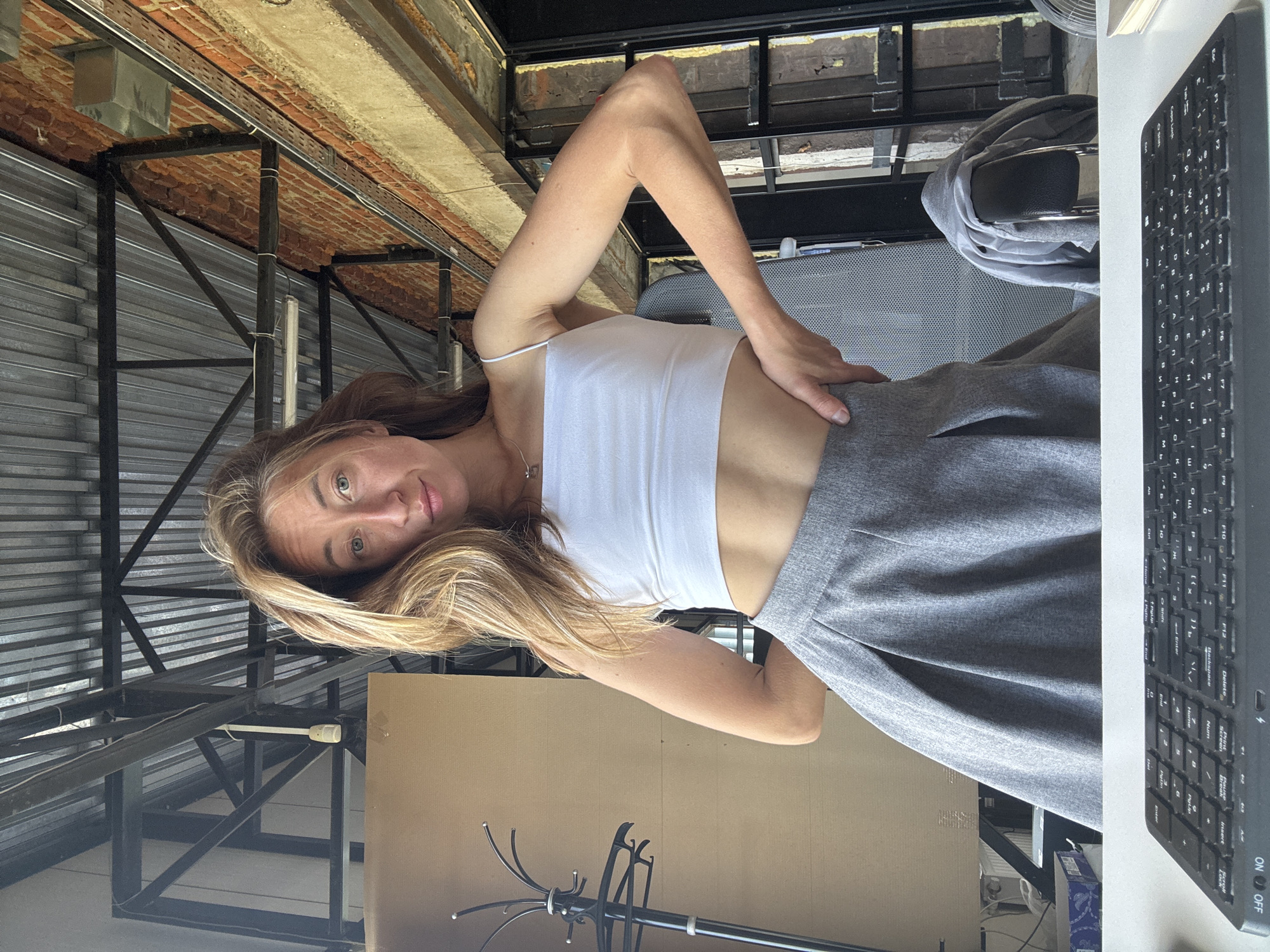 A woman with long, light brown hair is seated at a desk in what appears to be an industrial-style office space. She is wearing a white crop top and a grey pleated skirt, exposing her midriff. Her left hand is on her hip, and her right arm is bent at the elbow. The background features exposed brick walls, corrugated metal ceilings, and visible metal beams, suggesting a repurposed industrial building. A coat is draped over the back of a chair to her right. In the foreground, a black keyboard is visible on the desk. The lighting appears to be natural, coming from a large window or opening to the right, casting shadows and highlighting the textures of the environment. The overall impression is that of a casual, perhaps artistic or co-working, workspace. The image does not contain any specific visual cues to pinpoint a location within Chaikovsky, Russia.