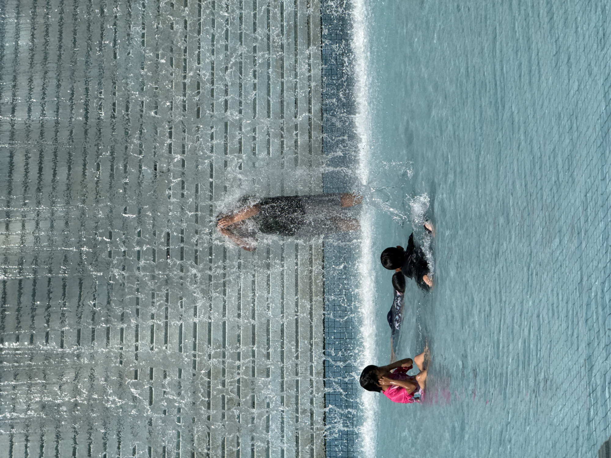 An overhead shot captures several children playing in a modern water feature in Kuala Lumpur, Malaysia, likely on a bright, sunny day. The image is oriented vertically, showing a textured wall on the left where water flows downwards like a waterfall, creating splashes and a shimmering effect. To the right, a shallow pool or water basin filled with light blue water is visible, with a grid-like pattern on its bottom, possibly from small tiles or a drain cover.
One child, wearing dark swimwear, is partially submerged near the base of the waterfall, actively splashing in the flowing water. Another child, wearing a bright pink top and patterned bottoms, is further into the pool, covering their face with their hands, possibly in response to splashes or the sunlight. A third child in patterned swimwear is partly visible between the two, also in the water. A fourth individual, potentially an older child or adult, is largely obscured by the heavy flow of water from the waterfall, wearing dark clothing.
The scene suggests a playful and refreshing activity, with the children enjoying the cool water amidst the dynamic flow of the waterfall. The smooth, flowing water contrasts with the textured wall and the subtle grid pattern of the pool floor. There is no visible text in the image.