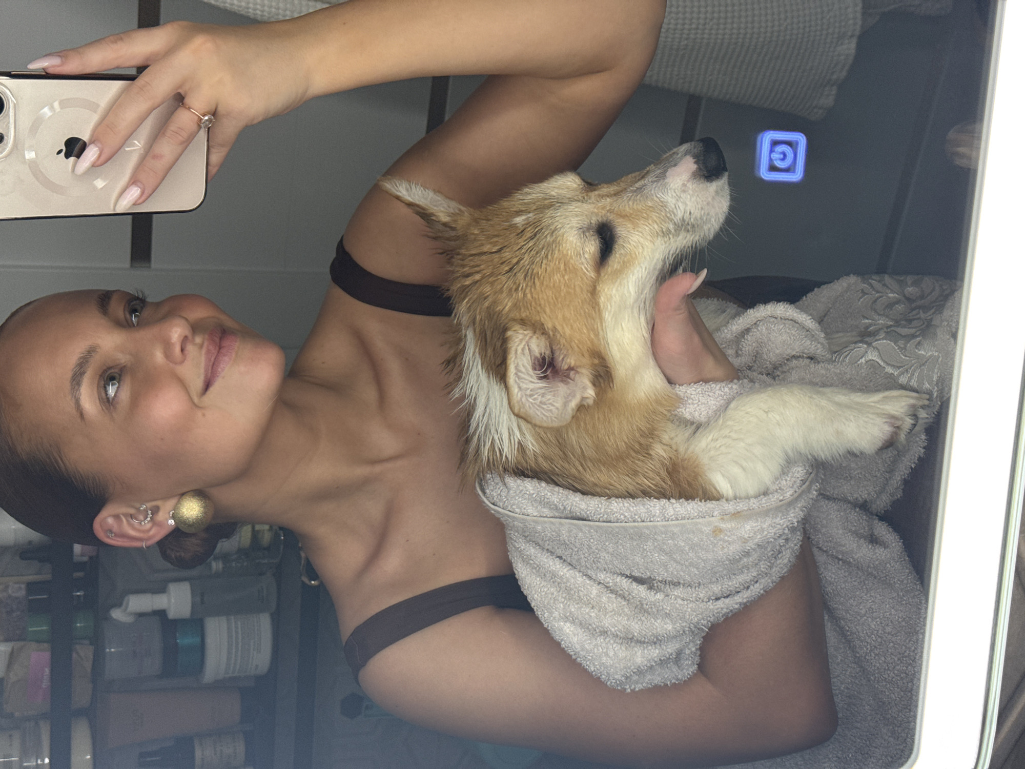 A woman is taking a selfie in a bathroom mirror while holding a Corgi dog wrapped in a towel. The woman has fair skin, light brown hair pulled back, and wears earrings. She is holding a mobile phone with an Apple logo. The dog appears to be wet. In the background, toiletries are visible on a shelf. A power button on the illuminated bathroom mirror is visible. The location is not discernible as being Chaikovsky, Russia from the given visual information.