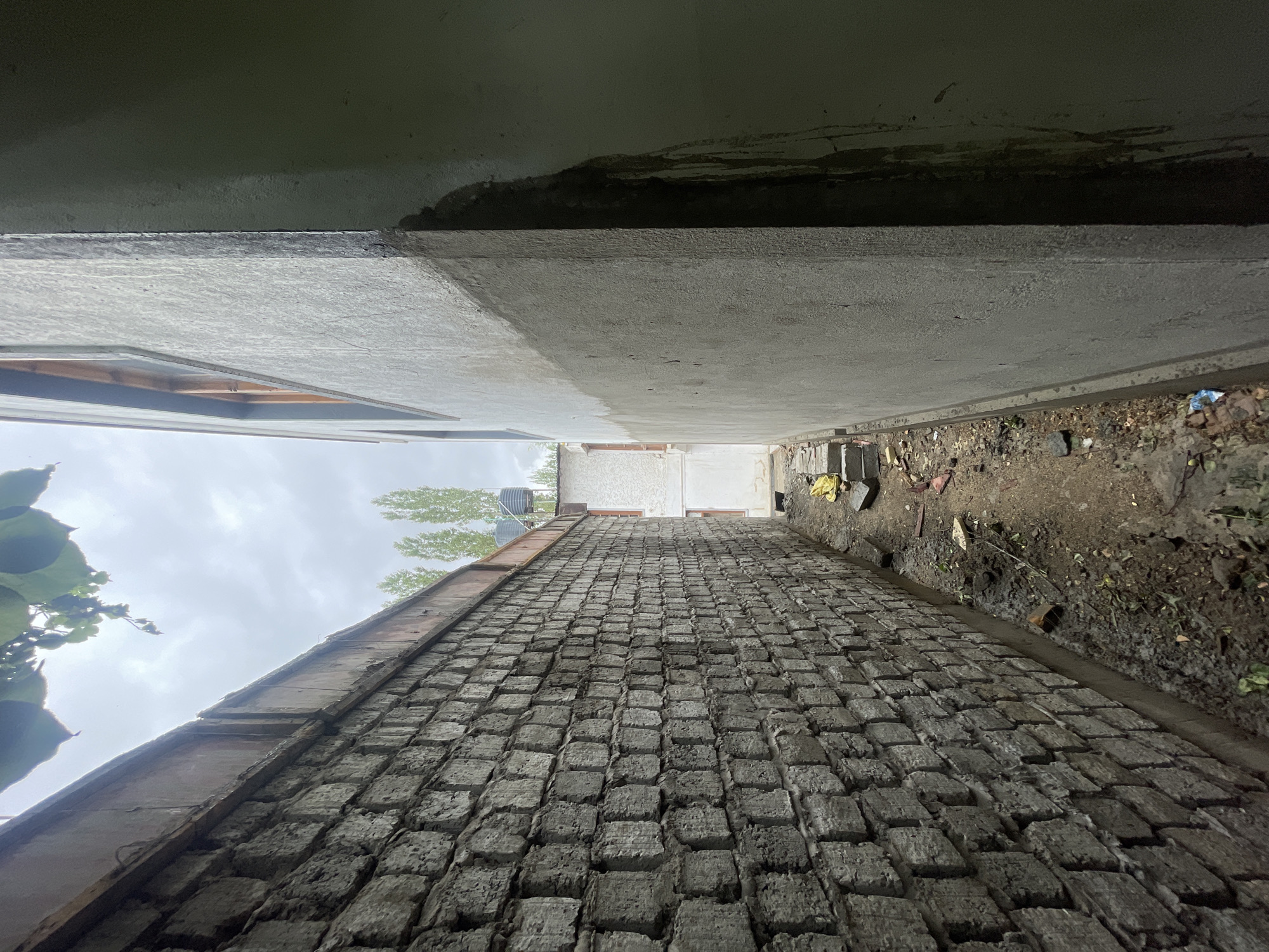 The image captures a narrow alleyway between two buildings in Leh, India. The left side of the alley is bordered by a textured, grey brick wall. The right side features a lighter-colored, plastered wall with visible water stains running down its surface. A wooden window frame is visible on the upper portion of the plastered wall. The ground of the alley is unpaved and littered with dirt, debris, and some construction materials, including yellow plastic sheeting and concrete blocks. In the background, more buildings and tall, slender trees are visible under a cloudy sky. The scene suggests an outdoor, possibly under-construction or neglected, urban environment in Leh.