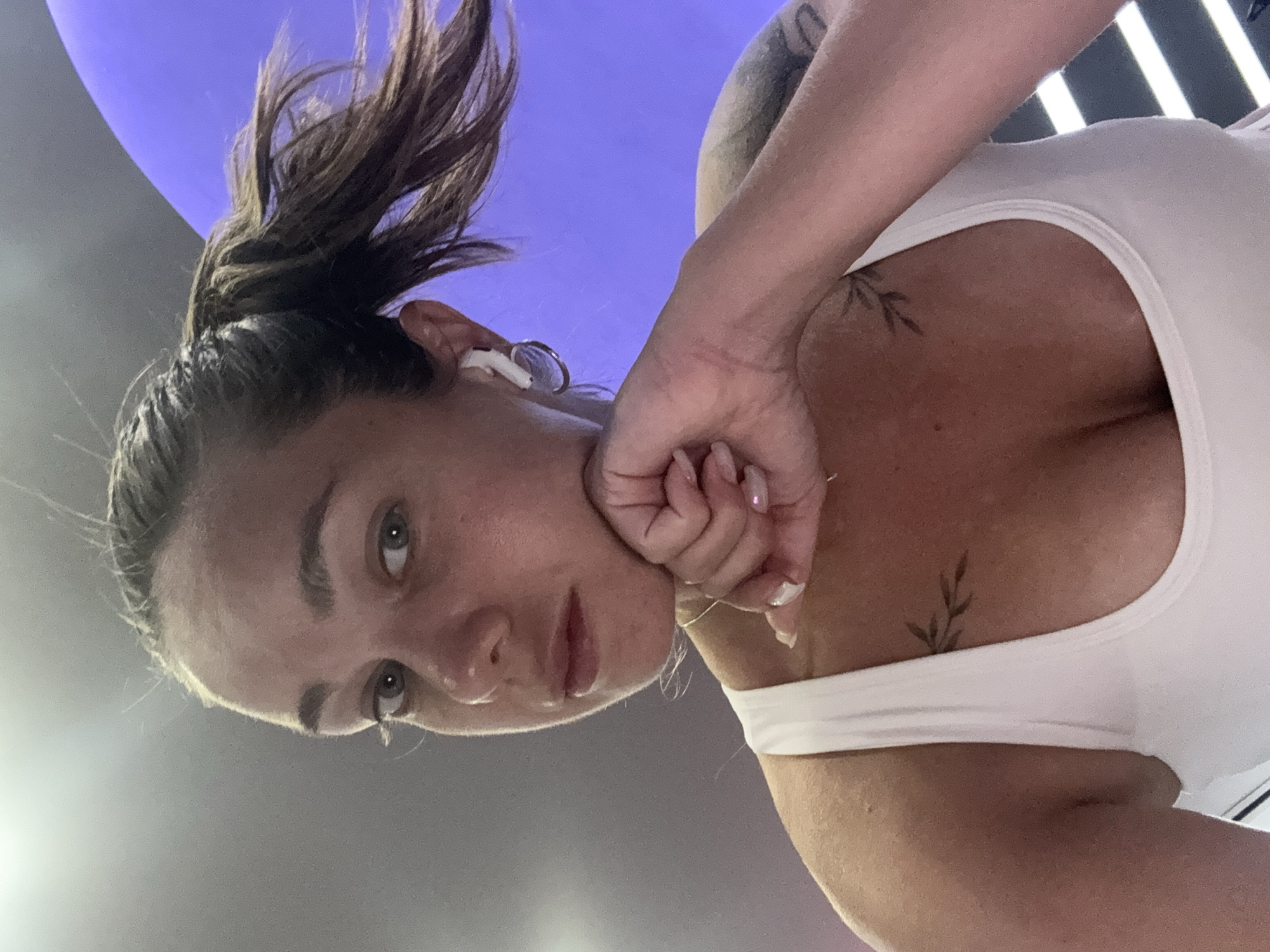 A woman with her hair in a ponytail, wearing a white tank top, is pictured from the chest up. She is resting her chin on her hand, which has manicured nails. She has a tattoo on her chest and another on her arm. She is wearing white earbuds and a thin necklace. The background is out of focus, with a purple shape on the right and a gray ceiling above. The lighting suggests an indoor setting. There is no visual information in the image to indicate the location in the city of Budva, Montenegro.