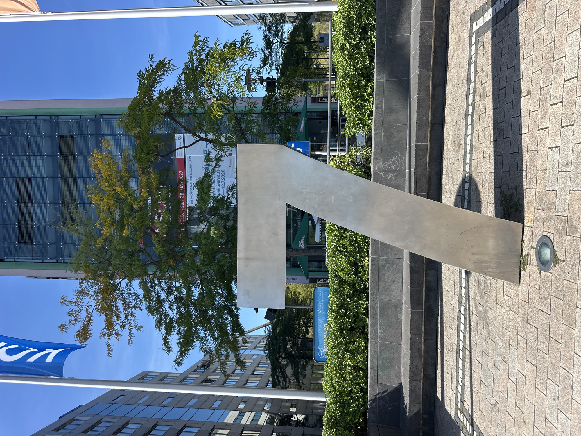 The image is a medium-shot photograph taken outdoors in Cologne, Germany, with a clear blue sky. In the foreground, a large, angled, metallic sculpture of the number "7" stands prominently. The sculpture is a light silver color and appears to be made of brushed metal. It is positioned on a paved area with light brown and grey square tiles. Behind the sculpture, a dark stone bench with a textured surface is visible, topped by a dense green hedge.
In the midground, the sculpture is silhouetted against a building with a facade primarily composed of glass panels. Green trees with yellowing leaves are situated in front of the building, partially obscuring it. A banner with text is also visible on the building, though it is not fully legible. A blue sign with white text is present near the hedge, also partially obscured by the foliage.
In the background, more modern buildings with glass facades and balconies line the street. A tall flagpole with a blue banner bearing a white "V" is visible on the left side of the frame, and another flagpole with an orange banner is on the right. Traffic lights and street lamps are also discernible among the trees and buildings. The overall impression is an urban outdoor setting, likely a public square or plaza in a commercial or business district of Cologne. There are no people visible in the frame.