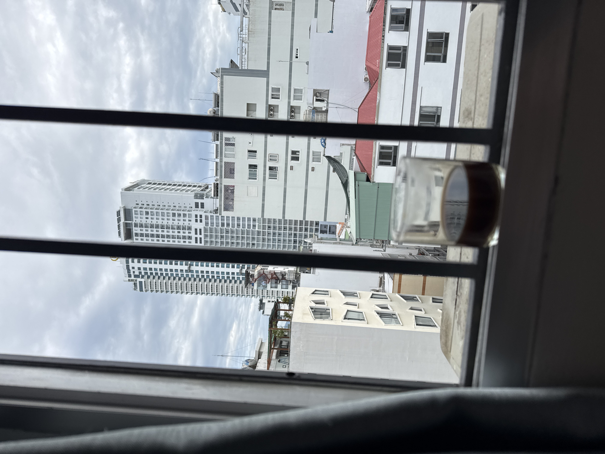 A shot taken from inside a building, looking out through a barred window at a city skyline. In the foreground, a glass of dark liquid, likely coffee, sits on a windowsill. The window bars are dark and prominent, partially obscuring the view.
Beyond the window, a cluster of buildings dominates the scene. The most striking is a tall, white skyscraper with numerous windows, a prominent structure in the urban landscape. To its left and right, other buildings of varying heights and architectural styles are visible, including some with white facades and others with red roofs. The sky above is overcast, with a cloudy, diffused light. The overall impression is a view of a dense urban environment, possibly in Nha Trang, Vietnam, given the architectural style and the common sight of skyscrapers and multi-story buildings in coastal Vietnamese cities. The scene captures a quiet moment, perhaps of someone enjoying a drink while observing the city.