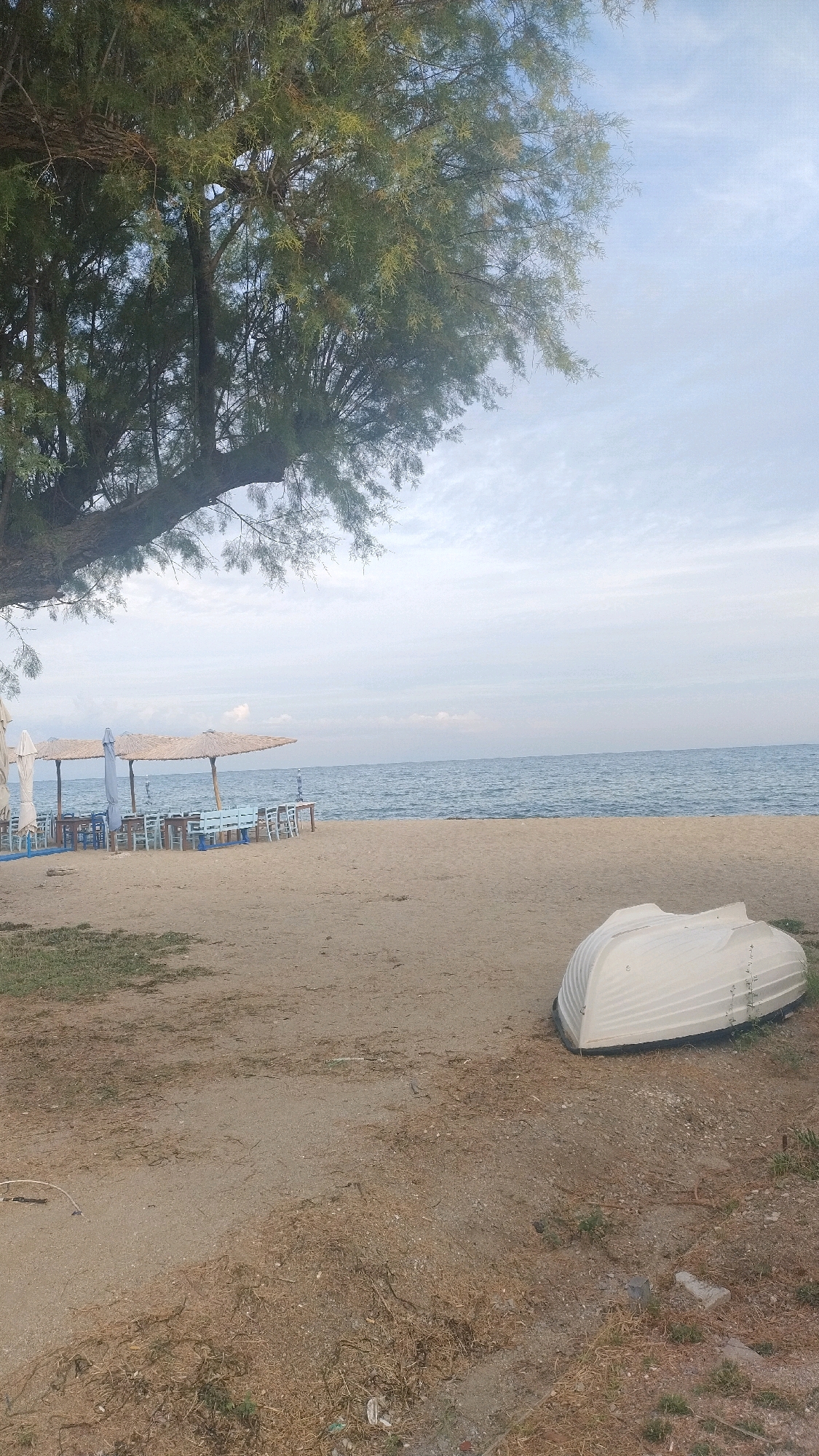 A white boat is overturned on the sandy shore of the Aegean Sea. The boat is in the foreground to the right of the image. Behind the boat is a sandy beach with some sparse vegetation. In the middle ground, there are several tables and chairs set up under thatched umbrellas, indicating a possible outdoor dining area. The sea stretches to the horizon under a cloudy sky. The image was taken in Neoi Epivates, Greece.