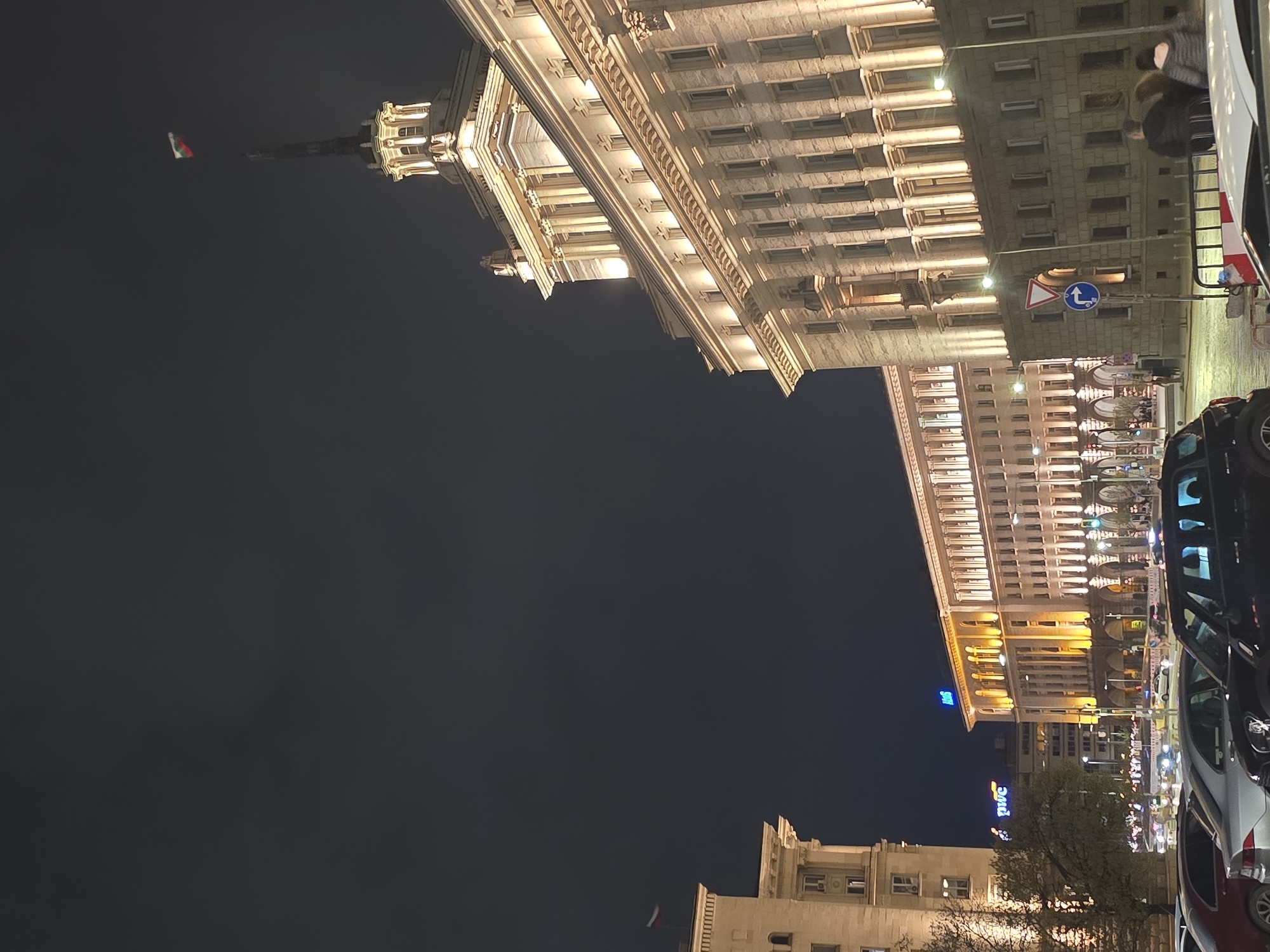 The image shows a night view in Sofia, Bulgaria, featuring illuminated buildings and street scenes. Dominating the frame are two large buildings, one brightly lit in beige tones and the other with similar architectural style but in yellow and orange hues. These appear to be government buildings in the city center.
A red, white, and green Bulgarian flag is visible on one of the buildings. Streetlights and building lights contribute to the overall illumination. The lower portion of the image shows other buildings, including one with a PwC sign.
On the right side of the image, cars are parked and moving, creating a sense of urban activity. A traffic sign is visible, indicating turning directions. People appear blurred in the street scene due to the night setting and the camera capturing their movement. The sky is dark, emphasizing the bright building facades.