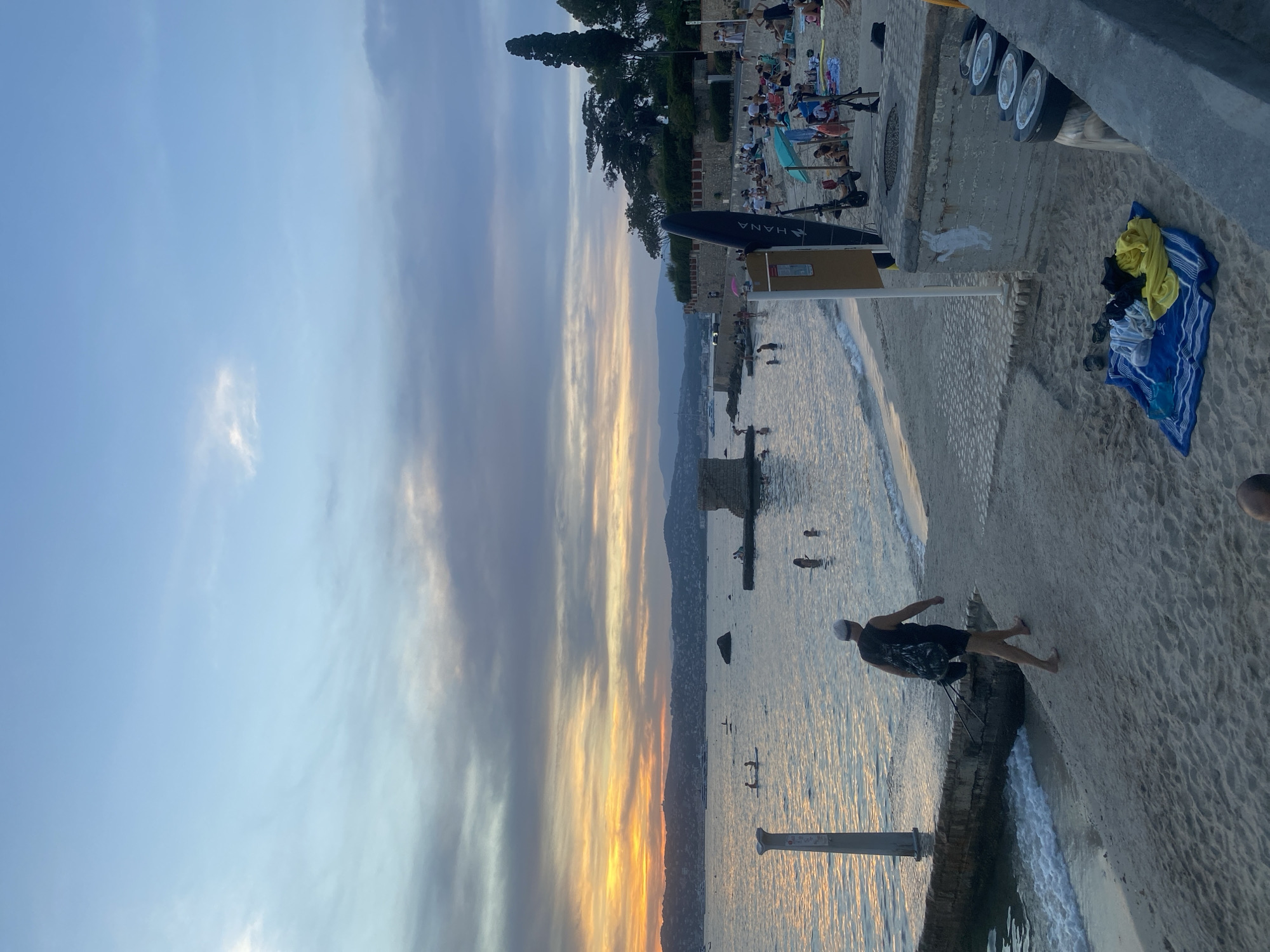 This is a medium shot image of a beach scene at sunset in Antibes, France. The sky displays a blend of warm orange and yellow hues from the setting sun, juxtaposed with streaks of gray and blue clouds. The sea is calm, reflecting the colors of the sky.
In the foreground, a person wearing a black tank top, black shorts, and a white cap is walking on the sand away from the water. They are carrying a backpack. Nearby, on the sand, a blue and white striped towel is spread out with various clothing items on top, including a yellow shirt, a black t-shirt, and striped pants. Several black circular objects, possibly trash cans, are lined up against a concrete structure.
The middle ground shows a sandy beach populated by many people. Some are lying on towels or under umbrellas, while others are in the shallow water. There are several stand-up paddleboards in the water. A dark-colored surfboard or paddleboard stands upright near a signpost. A fortified stone wall runs along the edge of the water, with a section of a ruin visible further down the coast.
The background features a coastline with rolling hills covered in greenery, leading to distant mountains. Yachts and boats are visible on the water in the distance. The overall impression is a peaceful evening at a popular beach.