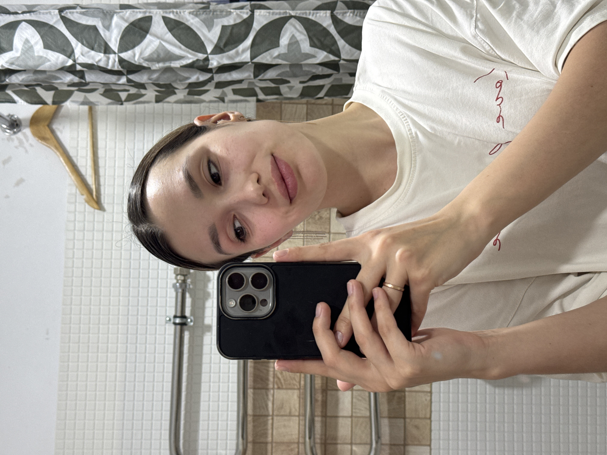 A young woman is captured taking a mirror selfie in what appears to be a bathroom. She holds a dark-colored smartphone with a prominent triple-lens camera system in her hands, reflecting her image. Her dark hair is pulled back, and she looks directly at the camera with a gentle, slight smile, showcasing natural facial features.
She is wearing a light, possibly off-white, short-sleeved t-shirt. On the chest area of her shirt, there is a red, stylized, handwritten-like text that is not fully decipherable but appears to be a cursive script. A simple gold-colored ring is visible on the ring finger of her right hand.
The background of the image features typical bathroom elements. A patterned shower curtain with a grey and white geometric design hangs in the upper portion. Below this, a white tiled wall with small square tiles extends downwards. A strip of darker, possibly brown, wainscoting or trim is visible near the bottom. Other visible objects include a wooden hanger or hook and metal plumbing fixtures, suggesting a functional bathroom space. The lighting is even, likely indoor and artificial.
