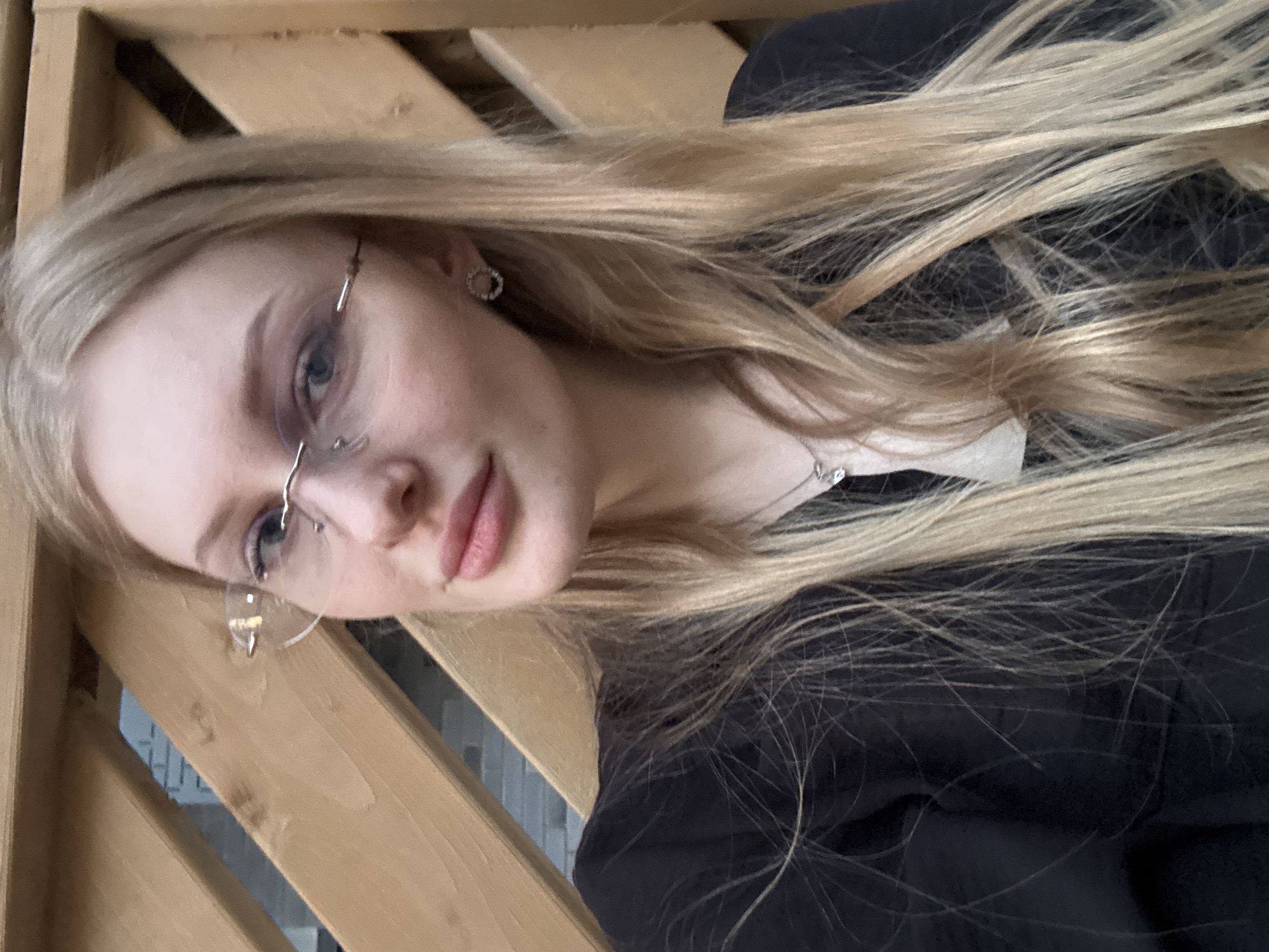 A young woman with light blonde hair and glasses is posing for a selfie. She is wearing a black jacket and a silver necklace. The background appears to be a wooden structure, possibly a fence or railing. While the location is given as Perm, Russia, there is insufficient information in the image to confirm this.