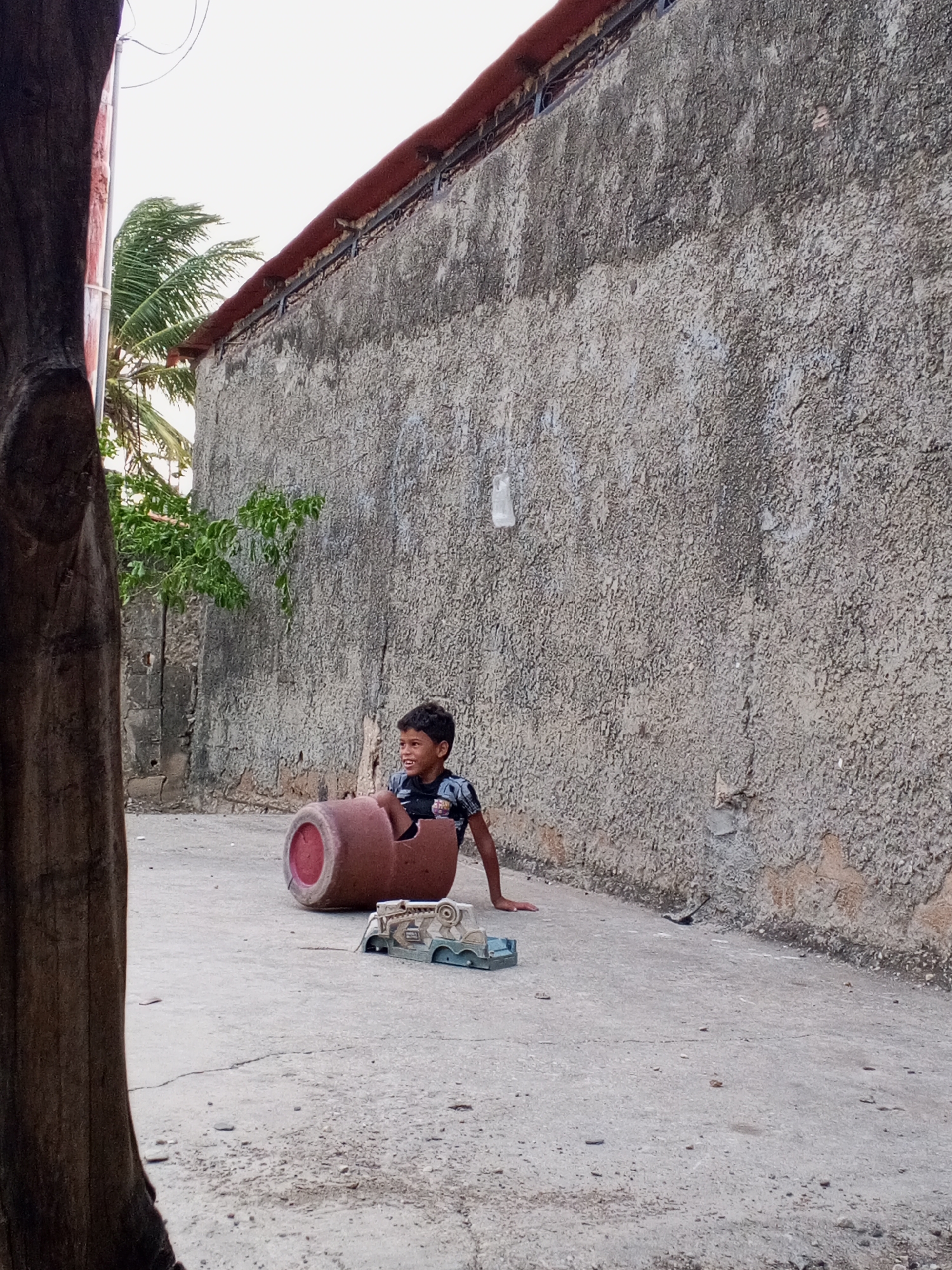 A young boy sits inside a broken clay pot on a concrete surface next to a toy truck. The scene is set outdoors, adjacent to a tall, textured concrete wall with a red roof overhang. To the left, a section of a tree trunk is visible, alongside a palm tree and other vegetation peeking over a shorter wall. The boy is looking off to the side and smiling.