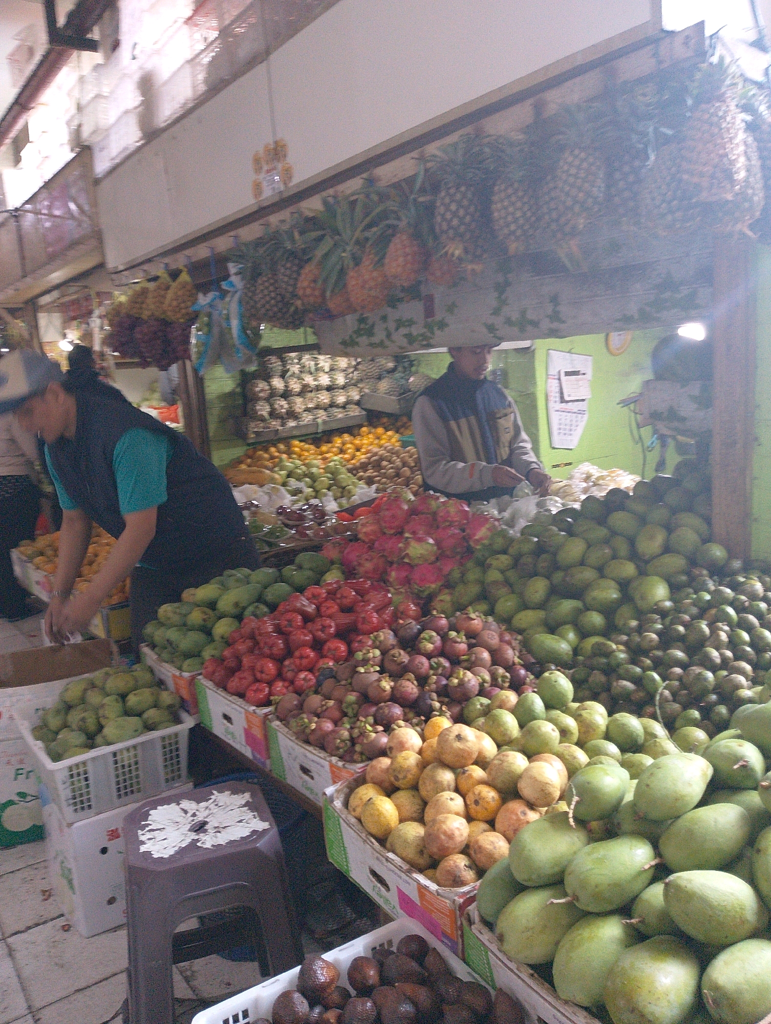The image shows a fruit stall in Lembang, Indonesia. A person wearing a dark vest over a turquoise shirt and a cap is seen arranging fruits. The stall is filled with a variety of fruits, including pineapples, guavas, mangosteens, and what appear to be rose apples. Various crates and baskets are used to display the produce. In the background, more fruits and possibly other market stalls are visible. The environment appears to be an open-air market or a covered market with natural light.