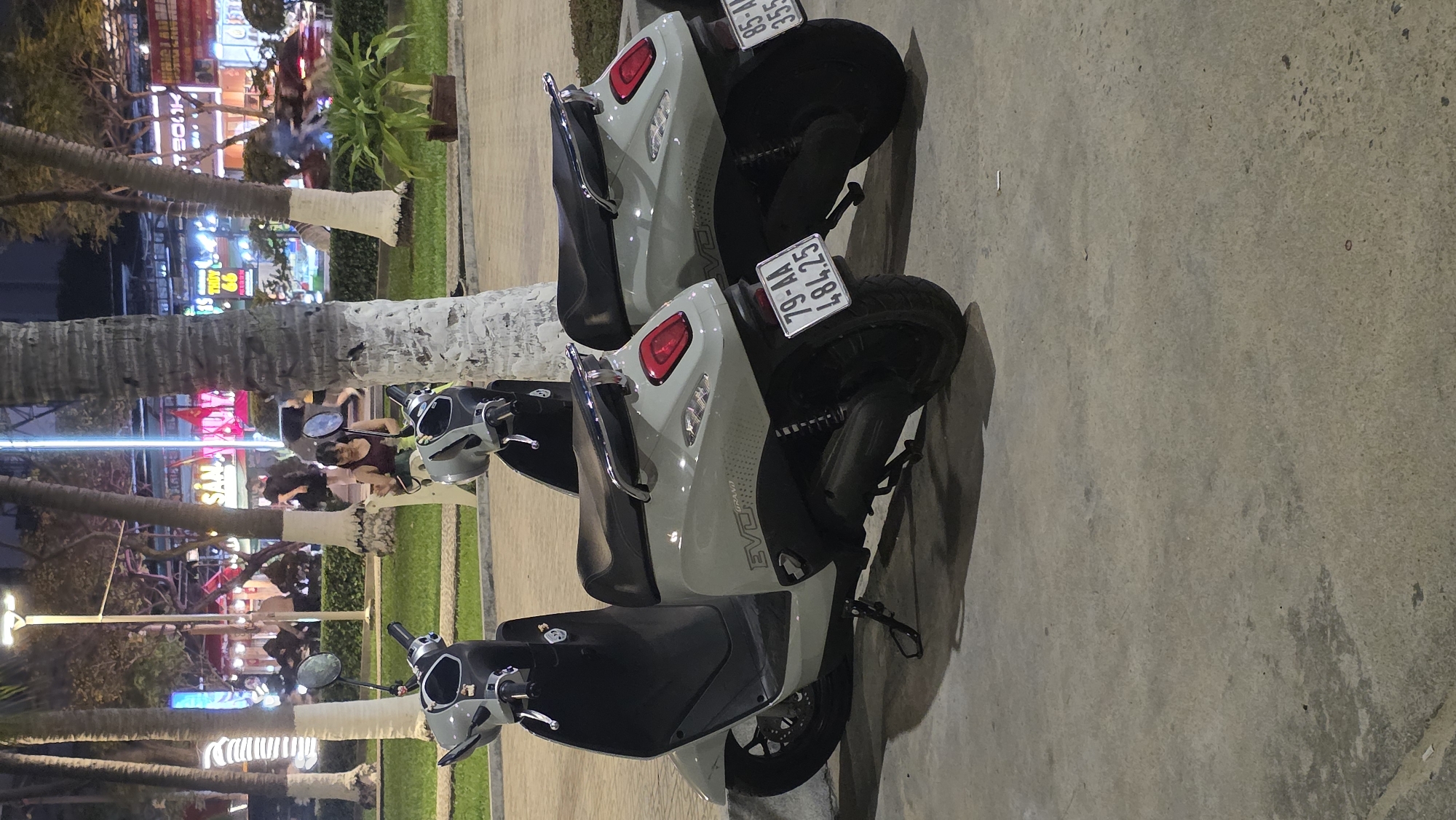 The image captures an evening or nighttime scene on a street, likely in an urban area like Nha Trang, Vietnam, given the architectural style and visible text. The foreground is dominated by two light grey electric scooters, identified by the "EVO" brand name on their side panels, parked on a concrete sidewalk. Both scooters feature black seats, chrome grab handles, and prominent red taillights. The license plate of the upper scooter reads "85-AA 145.58," while the lower scooter displays "79-AA 484.23."
Lining the sidewalk behind the scooters are several palm trees with white-painted bases, illuminated by artificial light sources. In the midground, between the trees and the background, several people are visible. One person in a dark top is near the front of the lower scooter, seemingly standing or adjusting something, while other figures are further back, suggesting a moderate level of pedestrian activity.
The background reveals a lively street with numerous brightly lit buildings, likely shops or restaurants, characterized by their vibrant signs and glowing facades. Visible Vietnamese text on these signs includes "SÁNG," "TÚN," and what appears to be "BOOK" (possibly "SOOK"). The overall setting is a bustling street scene after dark, with artificial lights casting reflections and shadows on the ground. The weather appears calm.