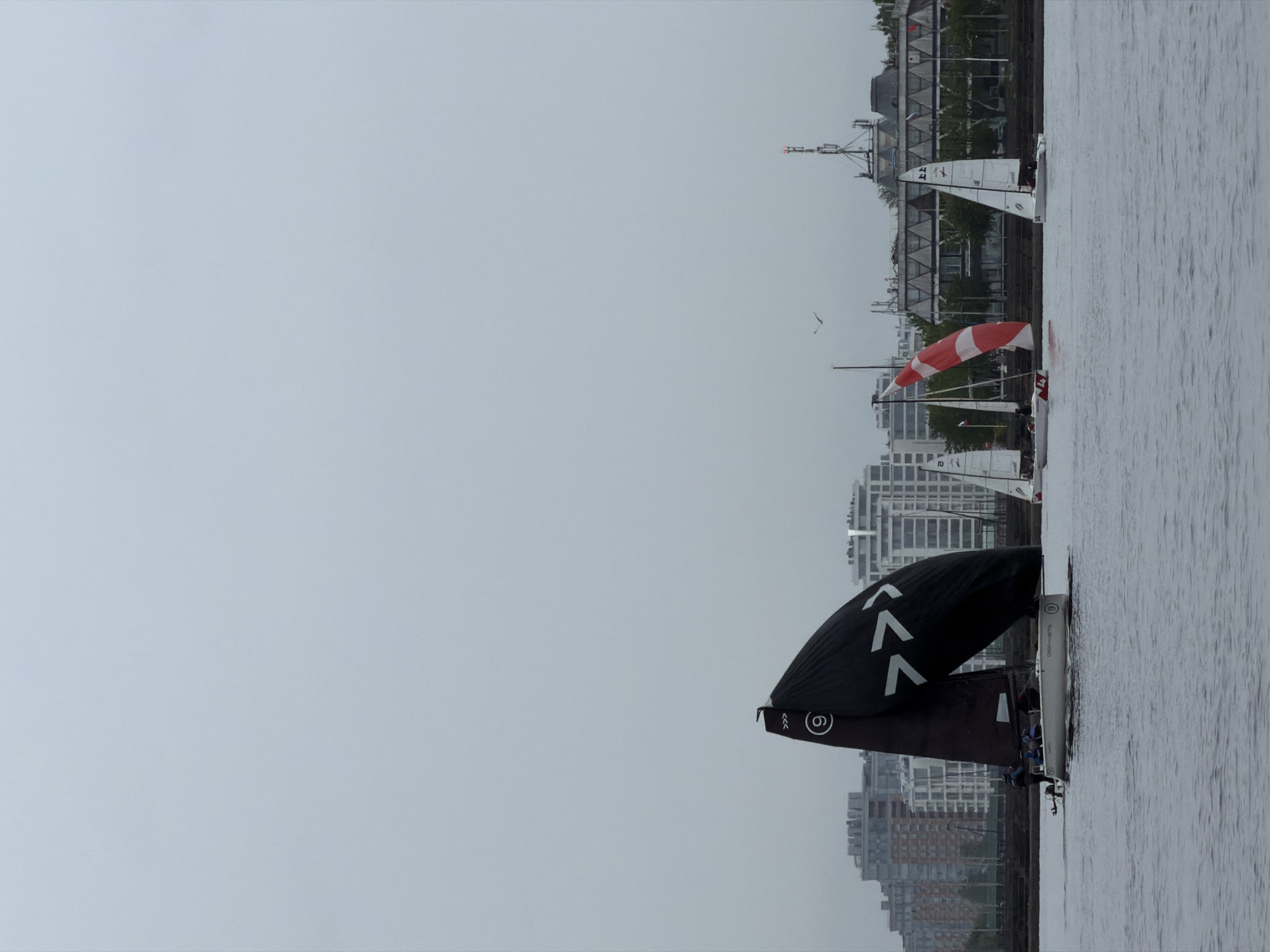 The image depicts a view of the water in what is determined to be the city of Saint Petersburg, Russia, evidenced by its distinctive urban architecture. Multiple sailboats populate the water, with one notably displaying a black sail marked with white arrows. Other sailboats exhibit sails in both white and red-and-white striped patterns. The backdrop consists of buildings and structures, indicative of an urban cityscape, juxtaposed against the water surface. The sky is overcast, contributing to the overall subdued and gray tone of the photograph.