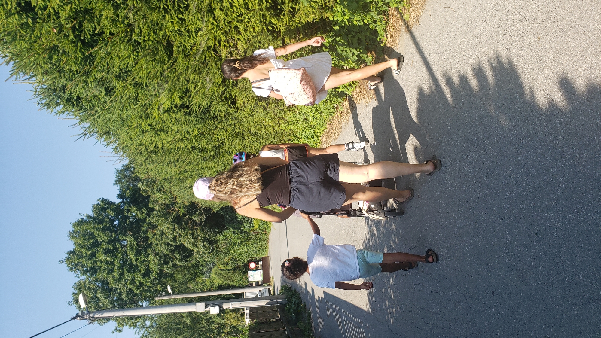 In Prešov, Slovakia, on a sunlit day, four individuals are walking on an asphalt path bordered by lush green trees. A woman in a black tank top and dark shorts, wearing a light pink cap, pushes a stroller. To her left, a young boy in a white t-shirt and light blue shorts walks ahead, holding a stroller handle. Behind the woman, a person on rollerblades, wearing a white tank top and dark shorts, is also moving forward. To the far right, a woman in a white dress and black flip-flops walks with a light-colored woven bag over her shoulder. The environment suggests a suburban or park-like setting, with a streetlamp visible in the distance and shadows cast by overhead foliage.