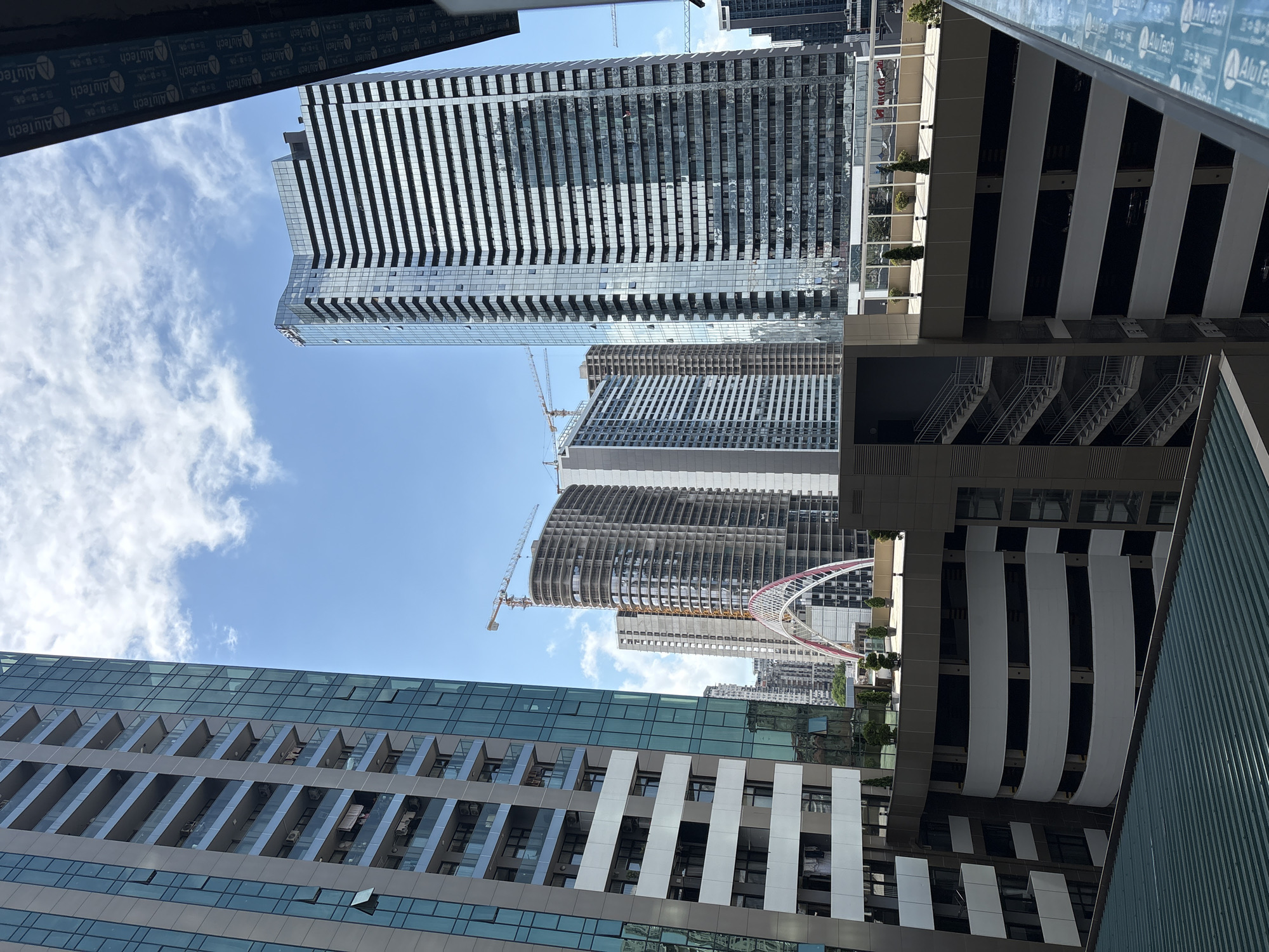 A view from a balcony looks out over a cityscape dominated by modern high-rise buildings and a partially constructed skyscraper. In the foreground, a green corrugated metal roof is visible, and to the right, a blue balcony railing with text "AluTech" repeated multiple times. Beyond the railing, a dark brown building rises, its facade featuring horizontal bands of windows.
The middle ground is occupied by a multi-story parking garage with a curving white facade and dark openings for parking spaces. A metal staircase is visible on the left side of the parking garage. On top of the parking garage, a flat roof area is visible with some small potted trees. Further back, a partially constructed skyscraper with an arched red and white pedestrian bridge in front of it is under construction. Cranes are visible above the construction site.
To the left, a tall residential building with glass balconies is visible, its facade characterized by alternating panels of white and glass. In the background, several other high-rise buildings of varying designs and heights fill the skyline under a bright blue sky with scattered white clouds. The image captures a dynamic urban environment with ongoing development.