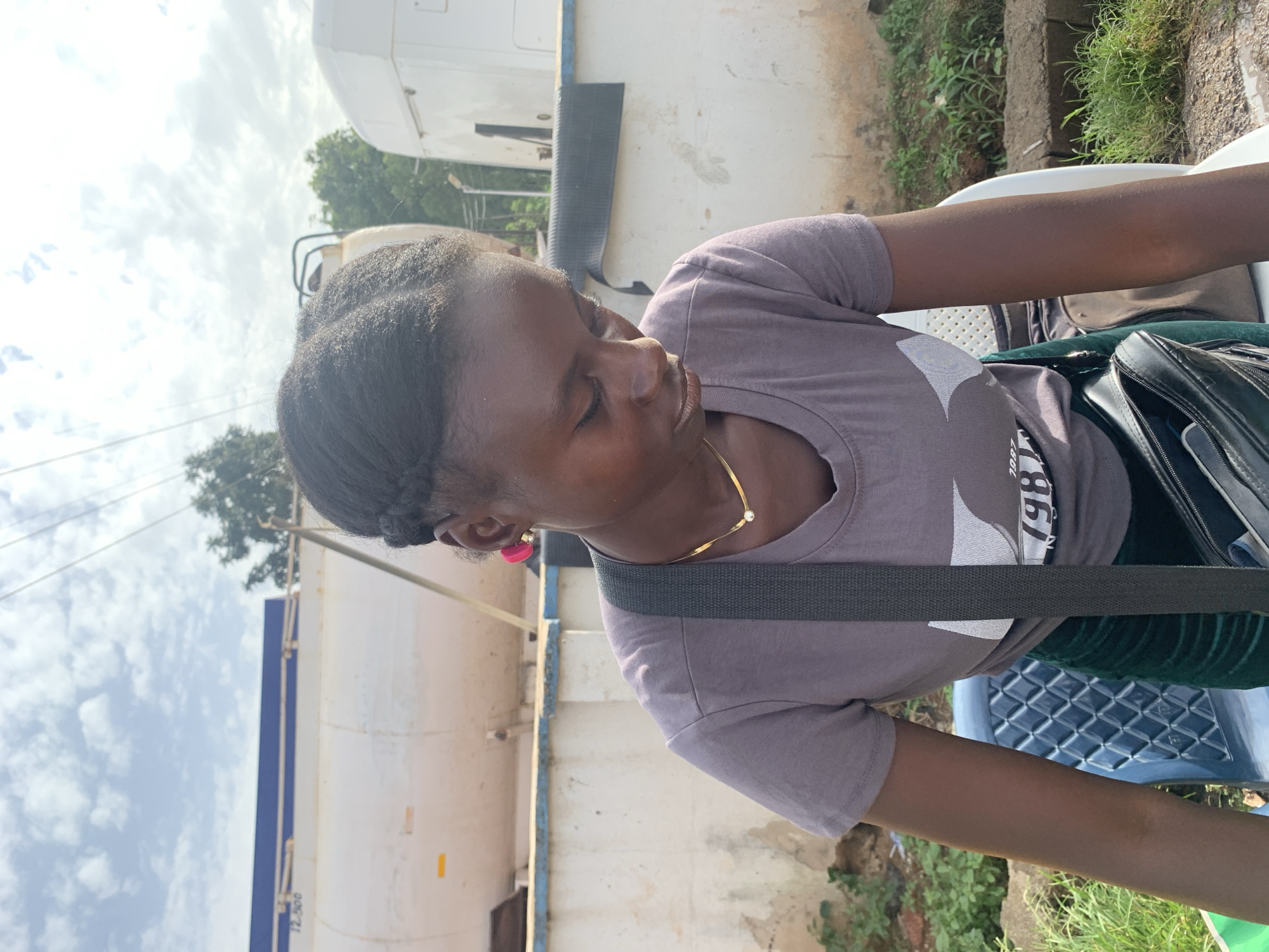 A young Black female with dark skin and braided hair is sitting outdoors, looking down. She is wearing a grey t-shirt with a graphic design and a dark green skirt. A gold necklace with a small pendant is visible around her neck, and she has a bright pink earring in her left ear. A dark grey strap from a bag crosses her chest. The background consists of a white wall, a white tanker truck with a blue stripe, and a white truck cab. Some trees and green vegetation are also present, as are power lines and a cloudy sky. The scene appears to be in an industrial or service area.
