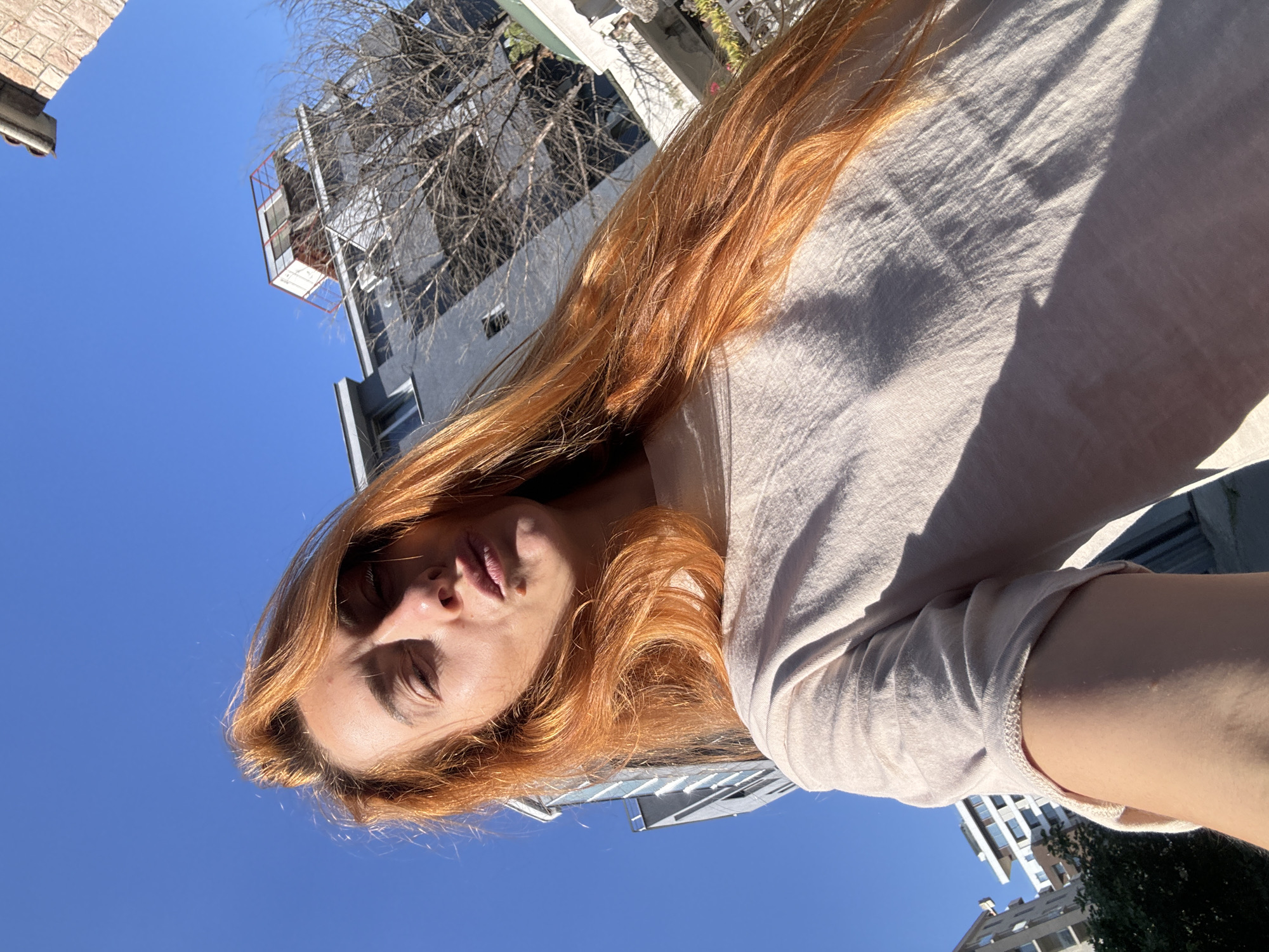 The image shows a selfie of a woman with long, reddish-blonde hair taken outdoors during the day. She is wearing a light-colored t-shirt. Her head is tilted, and she is making a kissing face with her eyes partially closed. The background consists of a clear blue sky and buildings, suggesting an urban environment. One building in the background appears to be modern with glass windows and balconies, while another has a more industrial or utilitarian structure on its rooftop. A bare tree is visible in front of one of the buildings. The lighting suggests sunlight casting shadows, with the sun appearing to be coming from the left of the frame. The overall impression is a casual snapshot taken outdoors. The location is identified as Budva, Montenegro.
