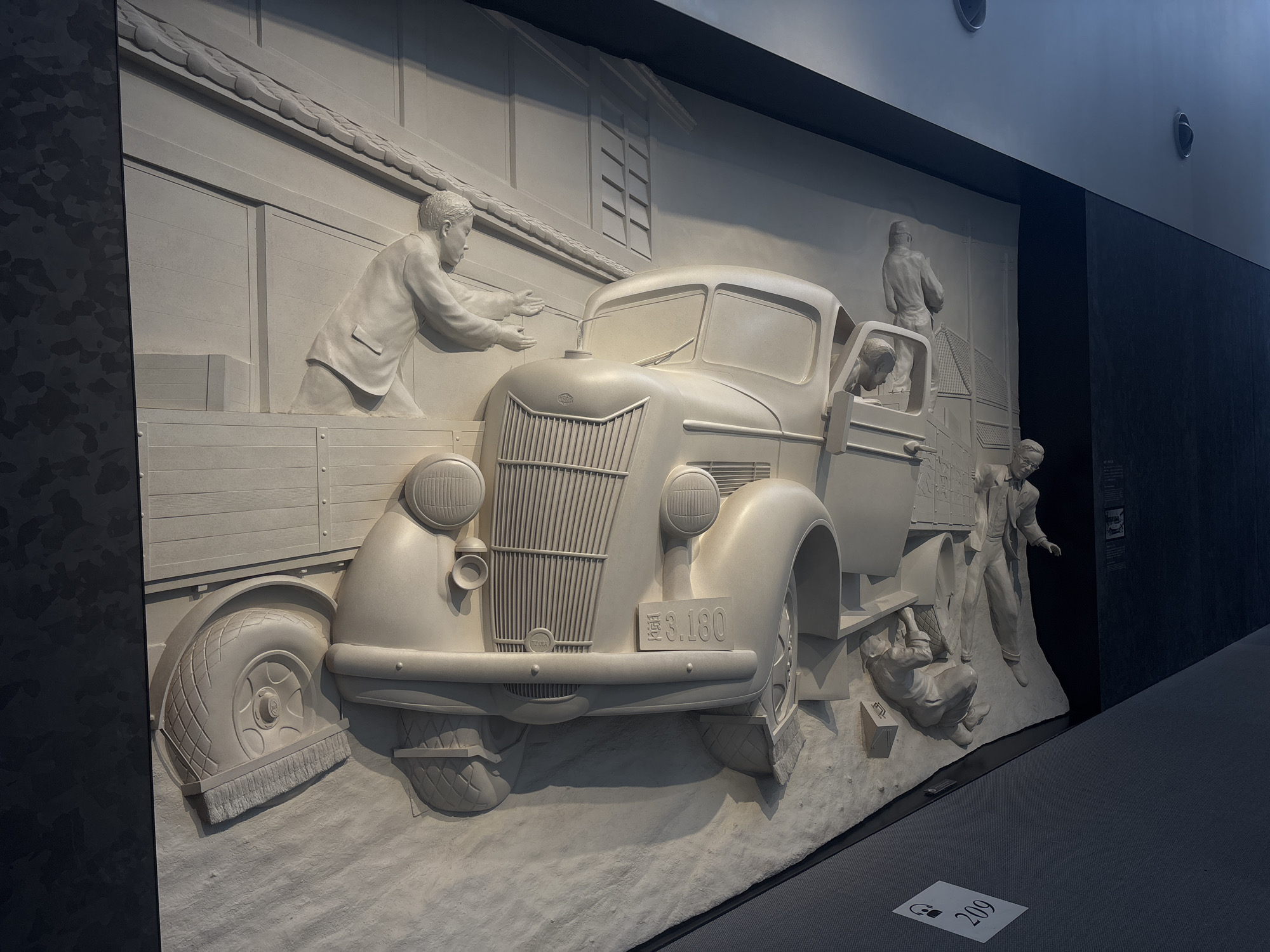 This image depicts a large, three-dimensional bas-relief sculpture, likely in a museum or exhibition space. The sculpture is rendered in a light-colored material, possibly plaster or stone, and is mounted on a dark wall. The scene portrays a historical moment involving a vintage truck and several figures.
The central focus is a light-colored truck, reminiscent of a mid-20th-century model, with a prominent grille and rounded headlights. A license plate on the front reads "S.3.180" in English and possibly Japanese characters. The truck has an open cab with figures inside and around it.
On the left side of the sculpture, a man in a suit is leaning over the truck's bed, appearing to be loading or unloading something. His hands are extended towards the truck.
In the truck's cab, a figure is visible, looking outwards. Another figure stands behind the truck, facing away from the viewer, possibly directing or overseeing the operation.
To the right of the truck, another man in a suit is leaning down, appearing to be working on the truck's tire or wheel, with a small suitcase next to him.
The background of the sculpture depicts architectural elements, possibly a building with a tiled roof and windows, suggesting an urban or industrial setting. The ground beneath the truck and figures is rendered in a textured, uneven manner, suggesting a natural or rough terrain.
The sculpture is illuminated by artificial lighting, casting shadows that enhance the three-dimensional effect and highlight the details of the figures and the vehicle. The overall presentation is clean and well-maintained, indicative of a public exhibition.
The provided hint that this is in Nagoya, Japan, suggests this sculpture might be related to the industrial history of the city, possibly commemorating transportation or early automotive development in Japan. The license plate format, with English characters and numbers, might be a stylistic choice to represent a historical period or a specific event.