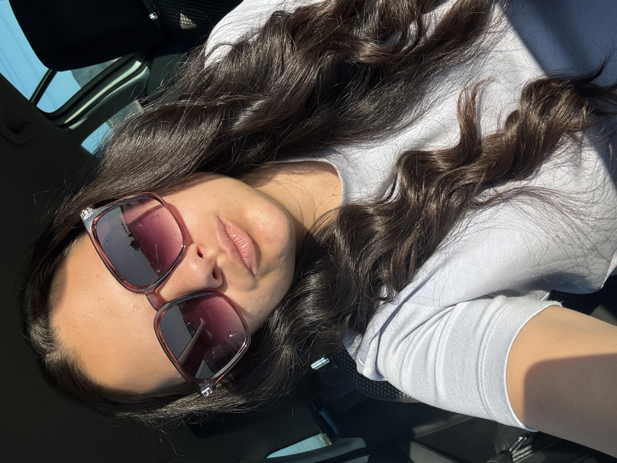 A woman with long, dark, wavy hair and sunglasses is sitting in a car. She is wearing a light blue t-shirt. The sun is shining, casting shadows on her face and hair. The background shows the interior of the car, including a headrest and a window. The image is taken from a close-up perspective, focusing on the woman's face and upper body. The scene is likely in Sliven, Bulgaria, given the context provided.