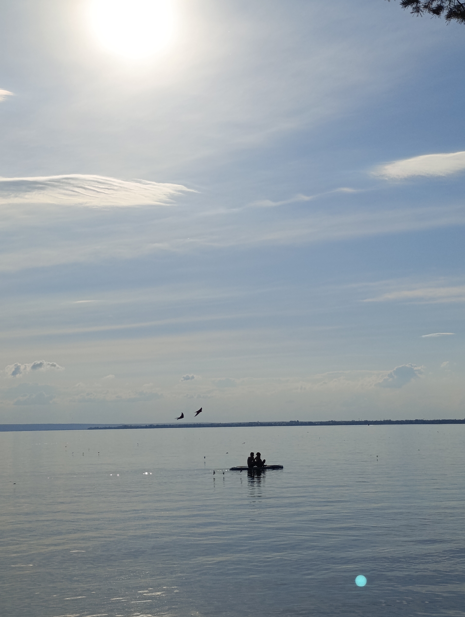 The image shows a serene lake scene with a bright sun in the upper left quadrant, casting a glare. In the middle of the lake, a dark silhouette of a paddleboard with two people on it is visible. Two dark bird silhouettes are flying in the upper middle section of the image. The horizon line is a faint strip of land with some indistinct structures. The water is calm and reflects the sky. The sky is partly cloudy with wispy white clouds.