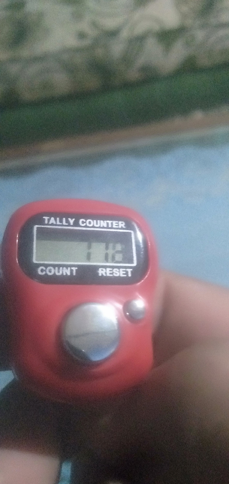 In the city of Plemahan, Indonesia, a red digital tally counter is held by a person. The counter displays the number 116. The counter has a screen that reads "TALLY COUNTER" and buttons labeled "COUNT" and "RESET". The background of the image is out of focus and appears to be a surface with a blue and green pattern.