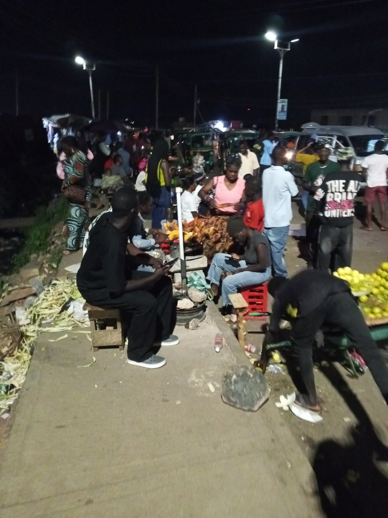 The image shows a bustling street scene at night in Bukuru, Nigeria. Several people are gathered near street vendors selling food and goods. Some individuals are seated, seemingly eating or interacting with the vendors. A man sits on a wooden stool using his phone. Another individual is bending down, possibly attending to merchandise. There is a display of lemons. There are streetlights illuminating the area. Vehicles, including possibly tuk-tuks, can be seen in the background. Trash and discarded corn husks litter the ground. The scene appears to be a typical night market setting in the city.