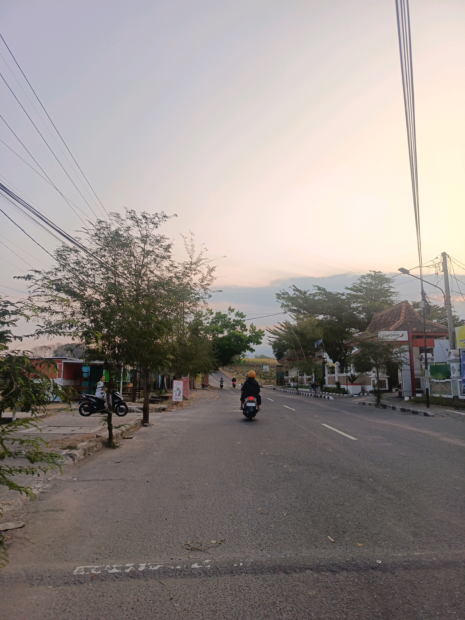 The image depicts a road scene in the city of Sumberagung, Indonesia, during what appears to be late afternoon or early evening, indicated by the soft, warm lighting and pale sky. The road is paved and has a white dashed line marking the center. On the left side of the road, there is a sidewalk with a few trees and some small buildings with red roofs and colorful facades. Several motorcycles are parked along the side of the road. One person on a motorcycle, wearing a yellow helmet and a dark jacket, is riding away from the viewer down the road. Further down the road on the right, there is a building with a distinct architectural style, featuring a tiled roof and an arched entrance with signage that includes "Bankjatim". More trees line the right side of the road, and there are streetlights and power lines overhead. Another motorcycle, with a rider wearing a white shirt, is visible on the left side. The overall impression is of a quiet, semi-urban environment.