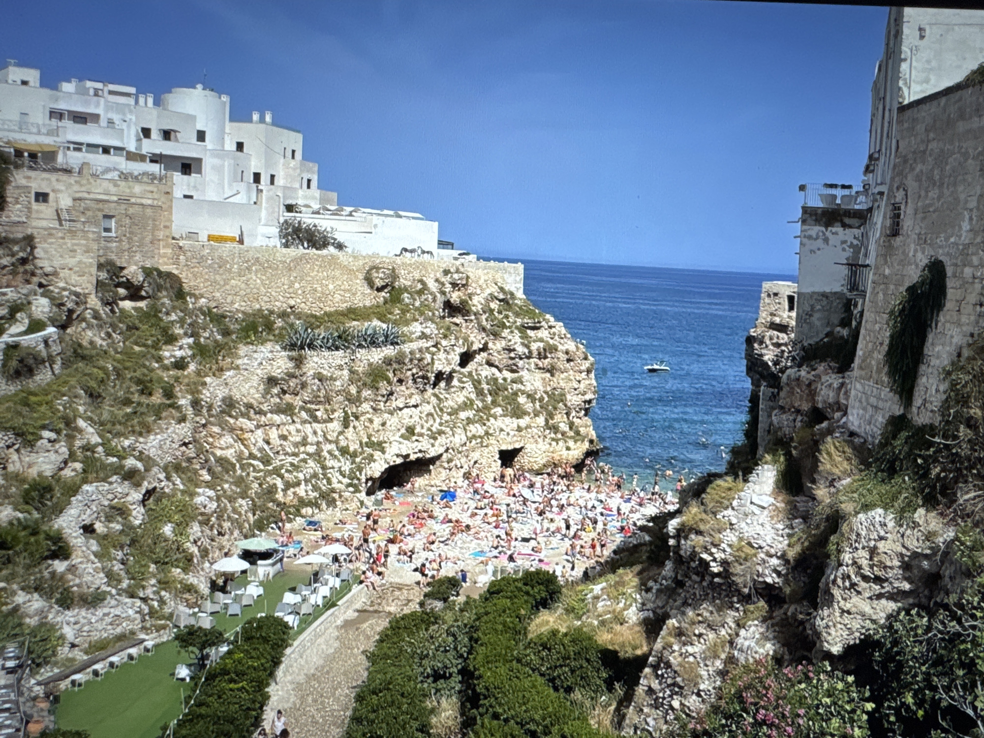 The image depicts a coastal scene with a beach crowded with people sunbathing and swimming. Buildings, some white and modern, others made of stone, are situated on cliffs overlooking the sea. A small white boat is visible in the turquoise water. Lush green vegetation grows on the rocky slopes. The sky is a clear, vibrant blue. The scene is a busy beach resort. This image is not of Porrentruy, Switzerland.