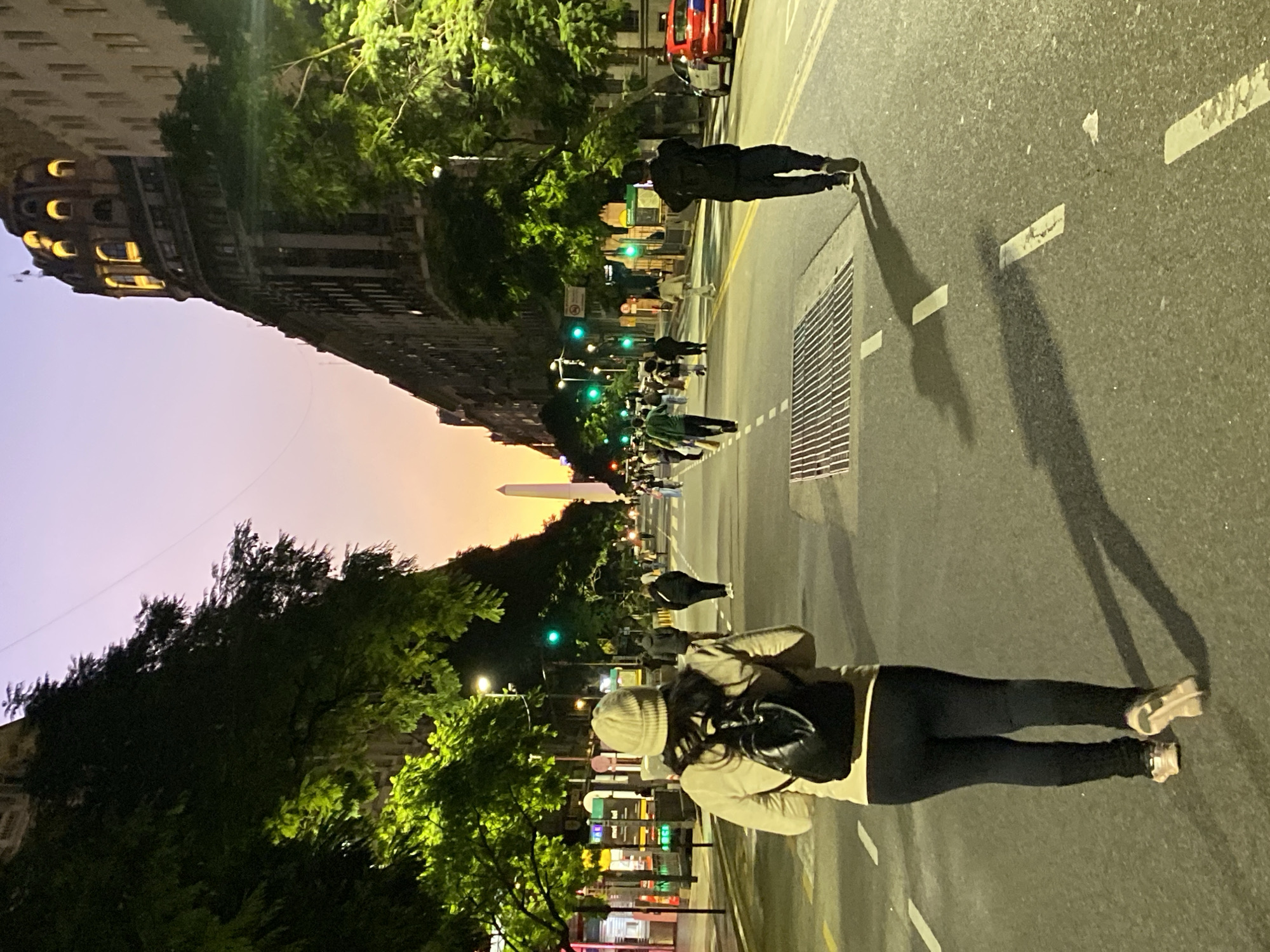 This image shows a street view in Buenos Aires, Argentina, with the Obelisco monument visible in the distance. The photograph is taken at street level. A woman with a backpack and wearing dark leggings, a beige coat, and a light-colored beanie is walking away from the camera. Other pedestrians are also walking in the same direction, with the Obelisco in the background. Buildings line the street with trees flanking both sides. Green traffic lights can be seen down the street. The sky is a mix of purple and orange. The street is illuminated by streetlights, casting shadows.