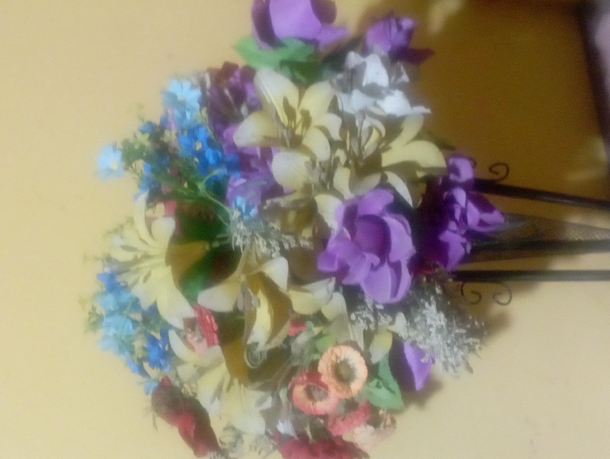The image shows a blurry, close-up shot of a bouquet of artificial flowers. The flowers are arranged in a vase that is partially visible at the bottom of the frame. The bouquet contains a variety of colors, including yellow, purple, blue, red, and white. The background is a plain, light yellow wall. The lighting is dim and appears to be artificial. There are no people or animals visible in the image. The scene is static and appears to be indoors. The exact location in Maos Kidul, Indonesia, cannot be determined from the image alone.