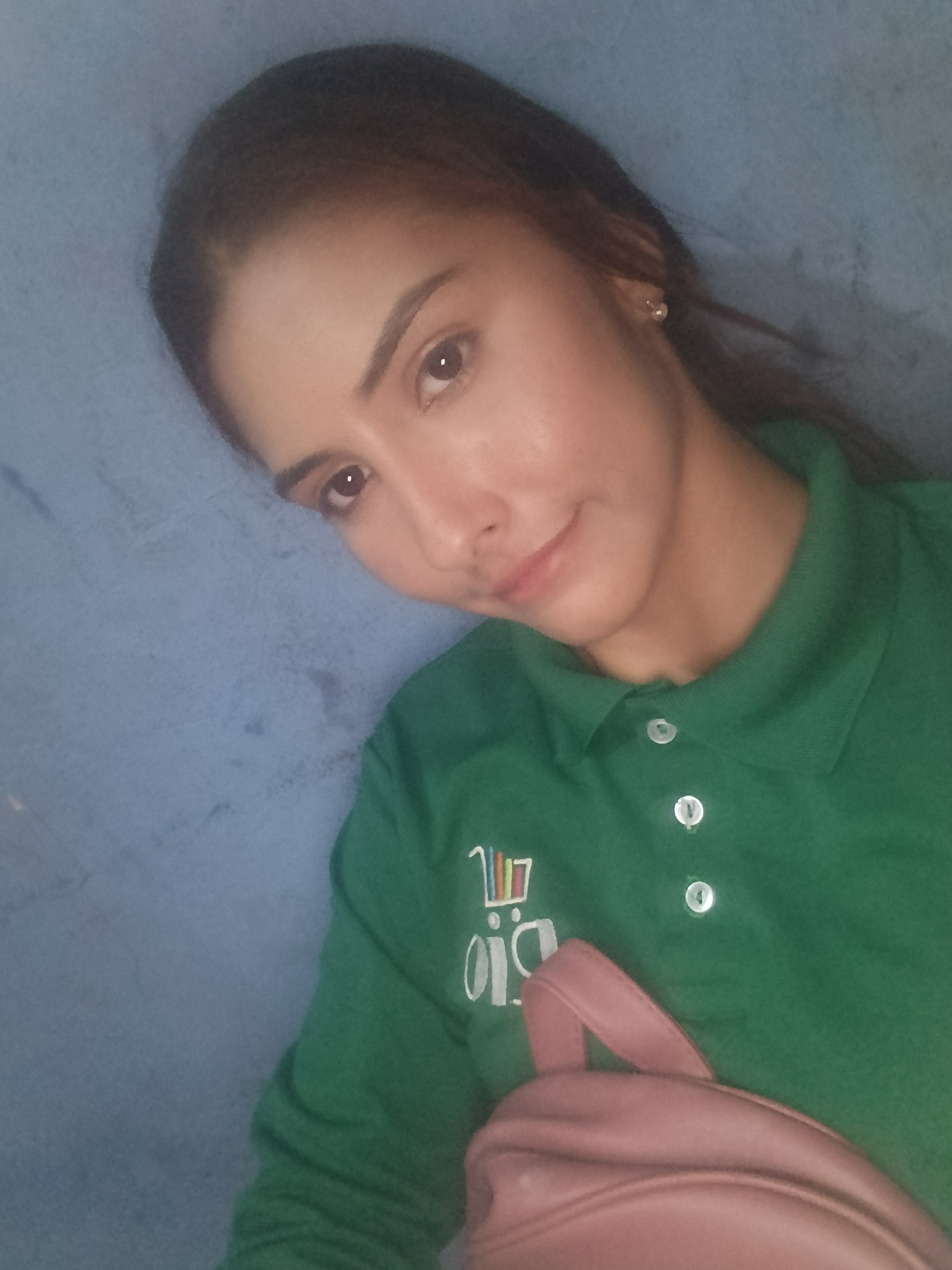 A woman with brown hair pulled back is wearing a green polo shirt with the word "BIO" and a shopping cart logo on the left side. She is carrying a pink backpack. The background is a textured blue wall. The location, Juan Griego, Venezuela, cannot be confirmed from the image itself.