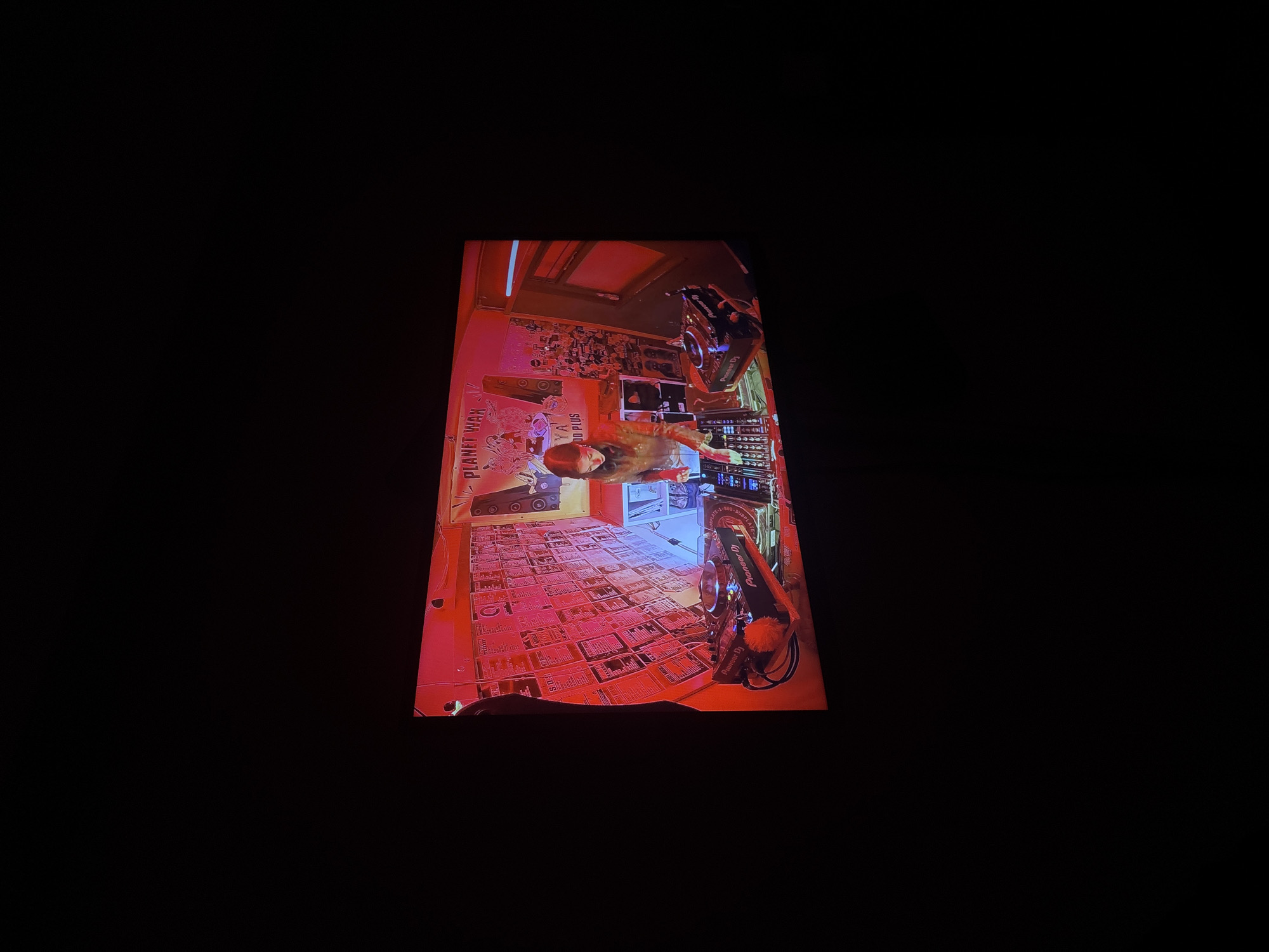 A vertically oriented screen displays a vibrant indoor scene, predominantly illuminated by warm red lighting. At the center, a woman with long hair stands behind a counter or DJ booth, possibly engaging with equipment. A large "Pioneer DJ" mixer/controller is prominent on the right side of the scene, clearly displaying its brand name.
Behind the woman, a wall appears to be covered with numerous records or album sleeves, creating a textured backdrop. To her left, part of a speaker system is visible. The floor in the foreground is densely arrayed with rectangular items, consistent with records, magazines, or flyers laid out in a grid. On the far left of the scene, the text "PLANET WAX" is visible vertically on a red sign or surface.
The setting strongly suggests a music-oriented venue, such as a club, bar, or record store, with the woman likely performing as a DJ. The red lighting creates an energetic and immersive ambiance. The original image captures this screen within a dark environment, possibly indicating it was taken at night or in a dimly lit room in Buenos Aires, Argentina.