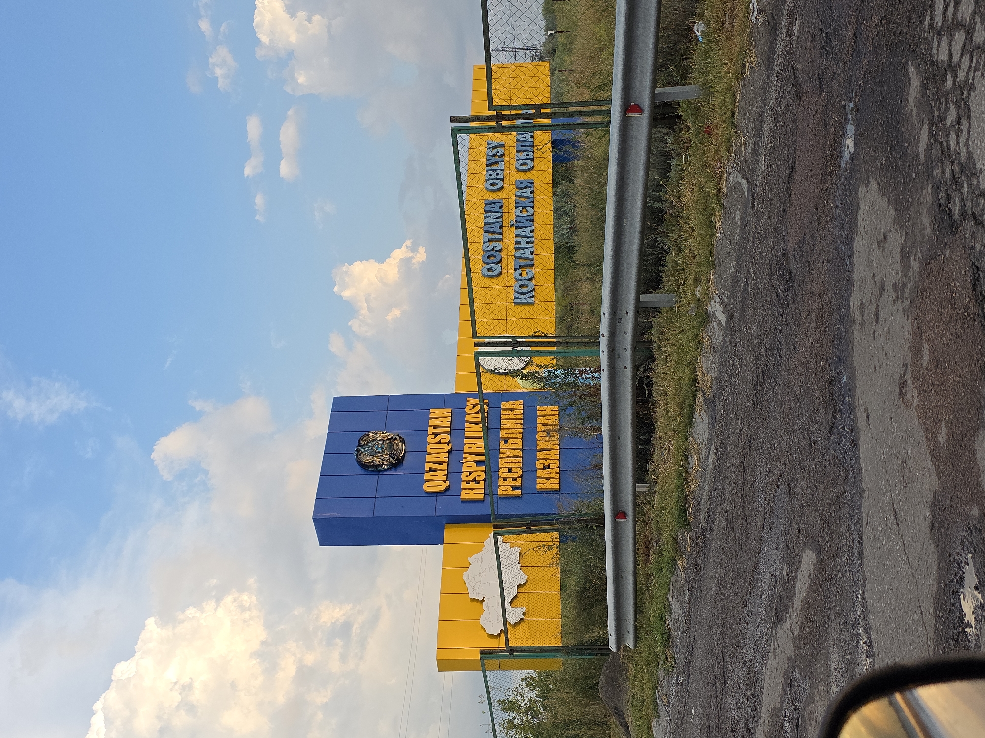 A wide-angle, eye-level, outdoor photograph captures a border crossing sign for Qostanay Region in Kazakhstan. The sign is predominantly blue and yellow. The blue section features the Kazakh coat of arms at the top and the words "QAZAQSTAN RESPYBLIKASY" in large yellow letters, followed by "РЕСПУБЛИКА" and "ҚАЗАҚСТАН" in smaller yellow letters. To the left of the blue section, a large yellow panel displays a white outline of Kazakhstan. To the right, another yellow panel has the words "QOSTANAI OBLYSY" and "КОСТАНАЙСКАЯ ОБЛАСТЬ" in large blue letters. A metal fence with a green frame runs across the foreground, with a metal guardrail above it. Beyond the fence, dry grasses and a few trees are visible. The sky is a bright blue with scattered white clouds. The road in the foreground is paved and shows signs of disrepair with potholes. The location is identified as a border crossing into the city of Podgorodka, Kazakhstan.