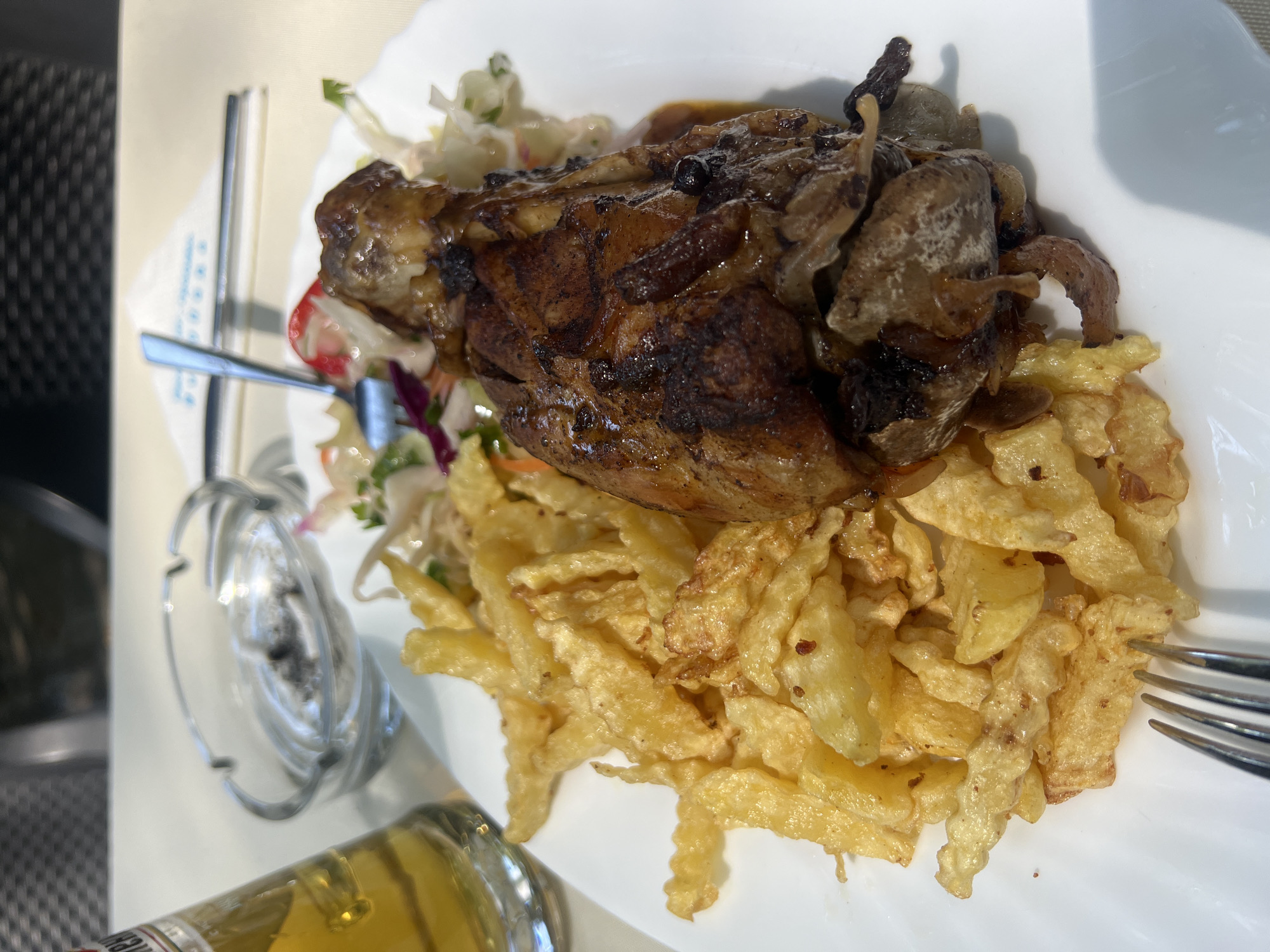A close-up, overhead shot captures a meal served on a white plate. The meal consists of a large piece of roasted meat, likely a pork knuckle, with crispy, golden-brown skin. It is accompanied by a generous portion of crinkle-cut French fries. A small side salad, appearing to be coleslaw with some red cabbage and a slice of radish, is situated behind the meat. To the left of the plate, a portion of a glass bottle containing a light-colored liquid, presumably beer, is visible. An ashtray with cigarette butts rests near the bottle. In the background, a fork is partially visible. The scene appears to be set outdoors on a table, suggested by the lighting and the blurry background elements that resemble outdoor seating. The image's perspective is from directly above the plate, focusing on the food. This meal was likely consumed in Bachkovo, Bulgaria, a location known for its monasteries and natural beauty, where such traditional Bulgarian cuisine might be offered at local eateries.