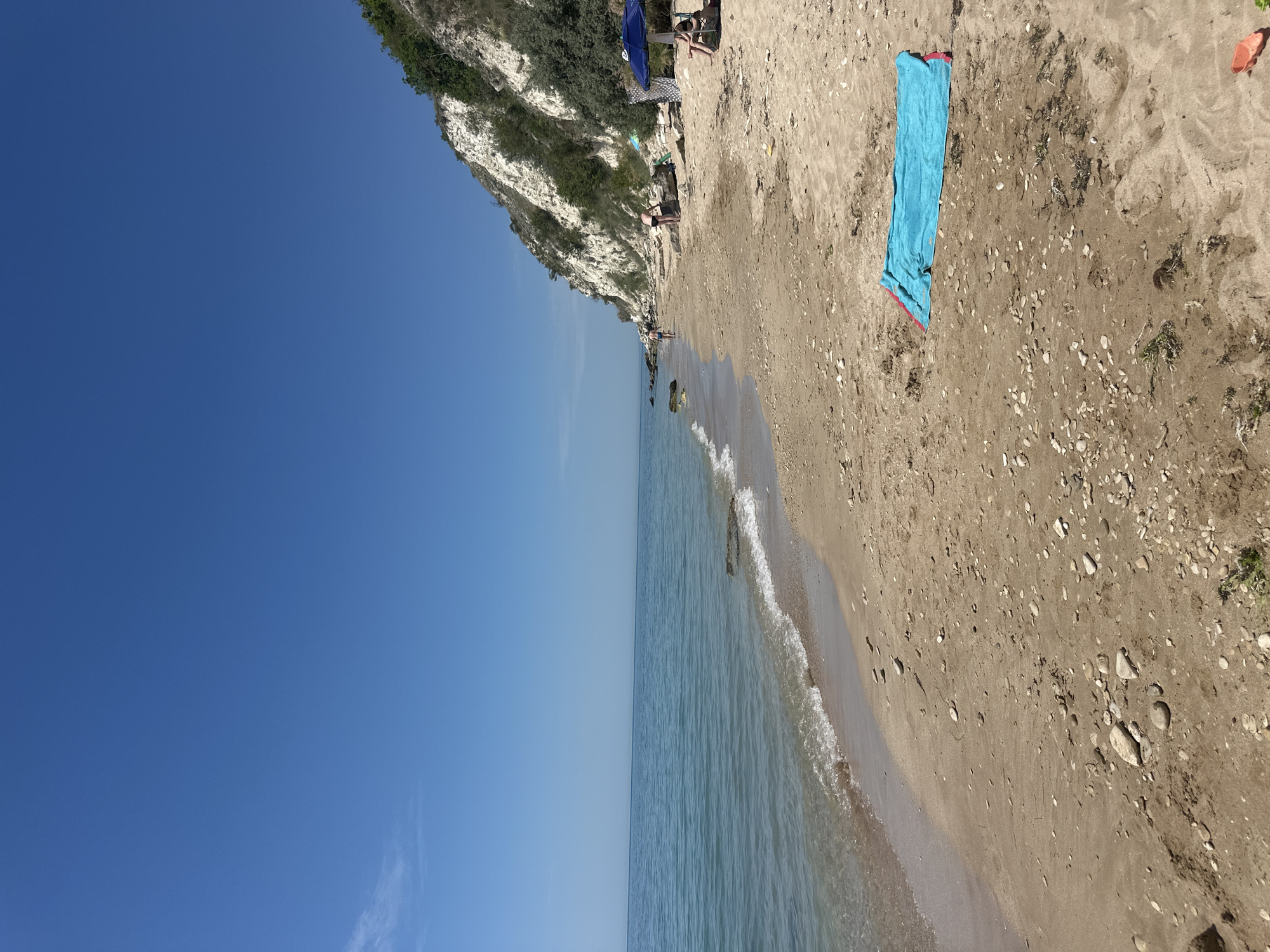 The image depicts a beach scene at Topola, Bulgaria, under a clear blue sky, suggesting a sunny day. The perspective is from a high angle looking down towards the shoreline. The beach is composed of light-colored sand interspersed with pebbles and some dried seaweed. To the left, a steep, rocky cliff face covered in green vegetation rises dramatically from the water's edge. The sea is calm and a light blue-green, with gentle waves lapping the shore.
On the sandy beach, a bright blue towel is laid out, suggesting someone is sunbathing or relaxing. In the background, near the cliff face and partially obscured by it, are a few people. One person is seated, and another appears to be standing. There is also a blue beach umbrella. The presence of people and the beach umbrella indicates recreational activity, likely on a summer day. The scene evokes a sense of peace and relaxation associated with a coastal vacation. No visible text is present in the image.