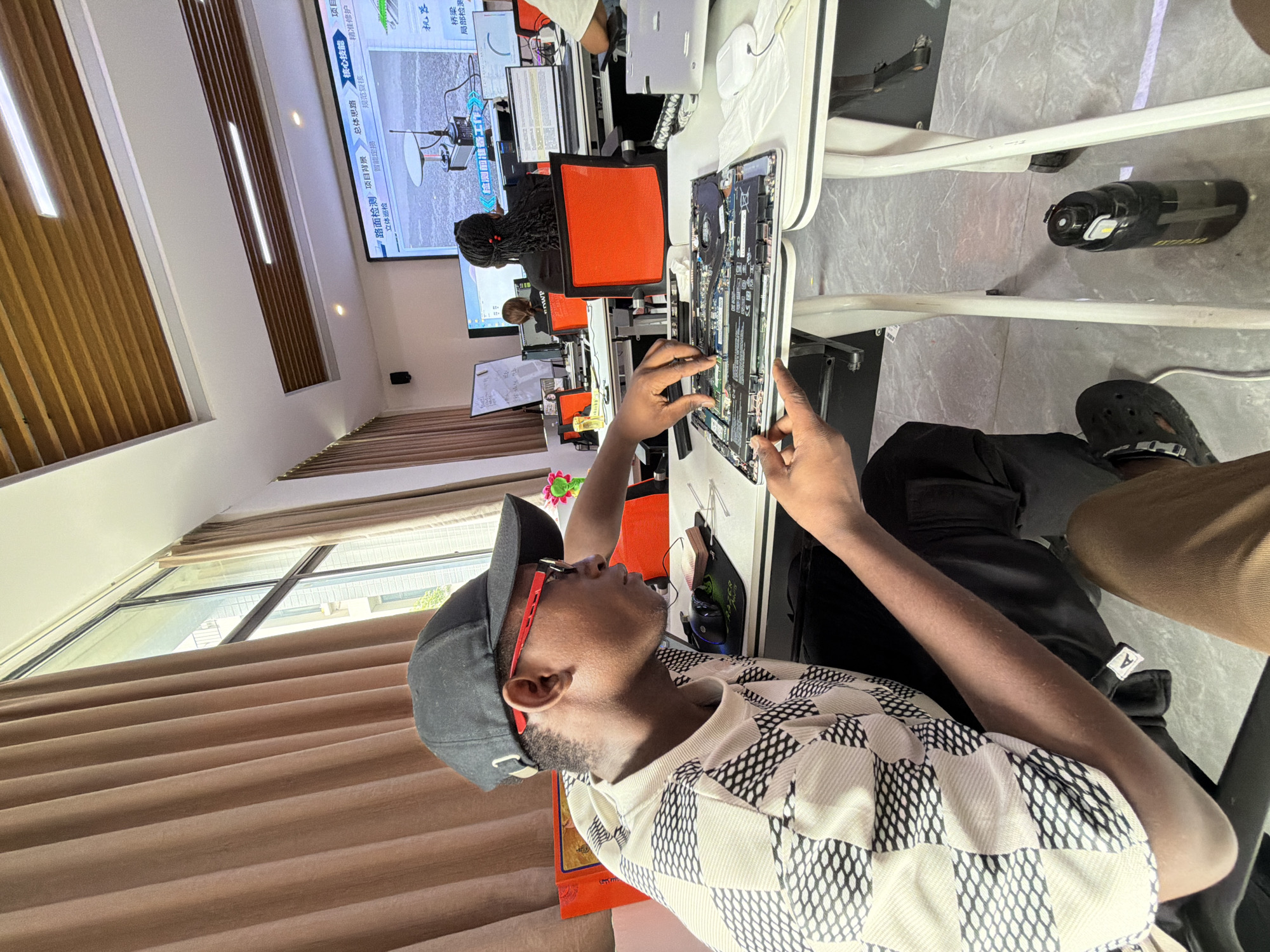 An overhead shot captures a young Black man wearing a black baseball cap, red-rimmed glasses, and a patterned t-shirt. He is leaning over a table, his hands working on the disassembled components of a laptop. The table is white, and a black computer mouse and a green mousepad are visible to his right. In the background, a large screen displays a presentation with Chinese text and an image of what appears to be a drone. Several other individuals are seated at tables, also engaged with computers and screens. The room is brightly lit, with natural light streaming in from a window covered by brown curtains. The floor is tiled in a light gray pattern. A black water bottle with a yellow label rests on the floor beneath the table. The setting appears to be an office or classroom environment within Jiangjin, China.