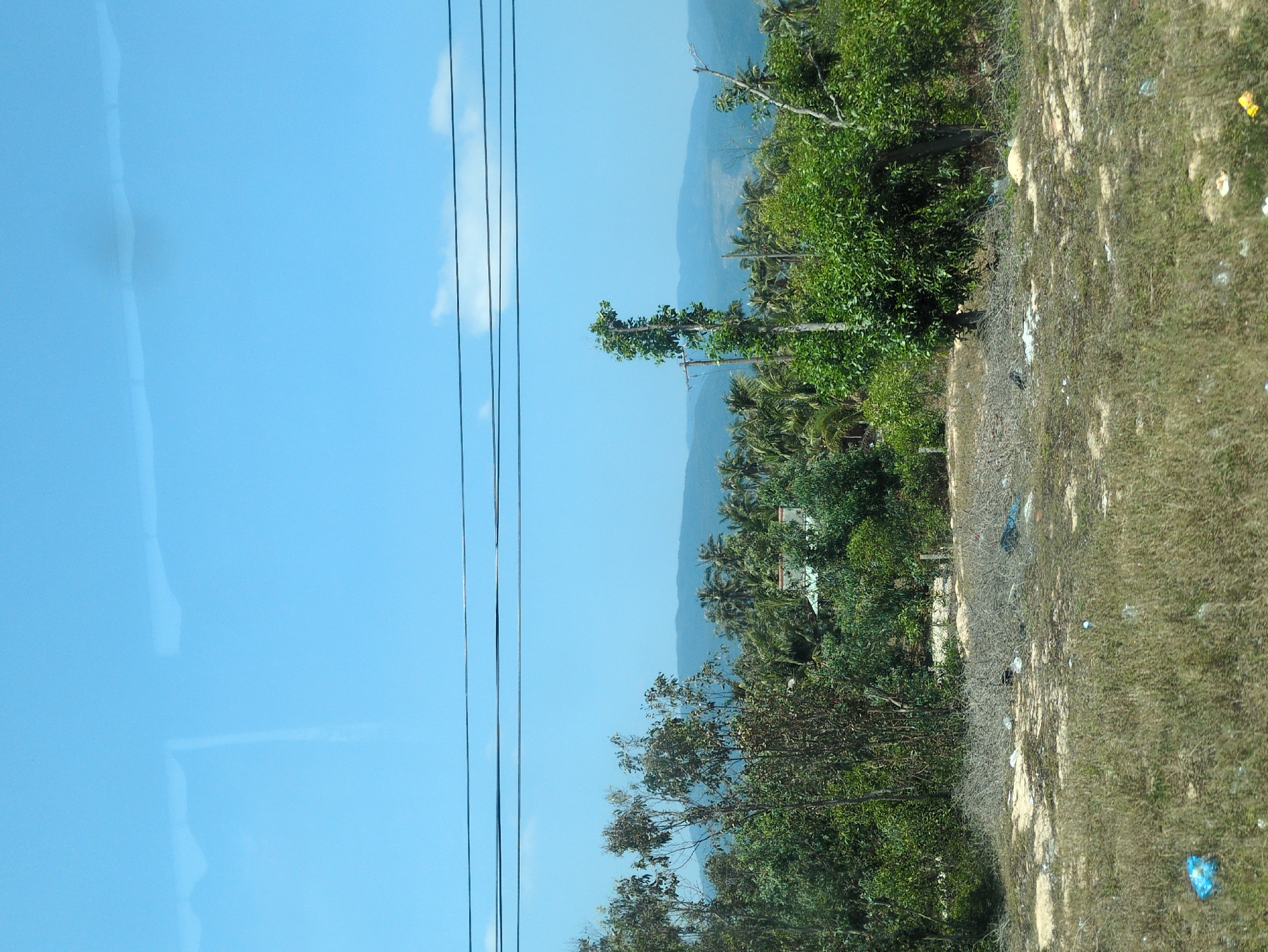 The image shows a view from inside a vehicle, looking out towards a landscape in Tuy Phong 4, Vietnam. On the left side of the image, there is a window with a clear blue reflection. Power lines run vertically in the middle ground. Beyond the power lines, there is a landscape with lush green vegetation, including trees and possibly coconut palms, against a backdrop of distant blue mountains. The foreground on the right side is a patch of dry, grassy land. There is some trash visible on the ground.