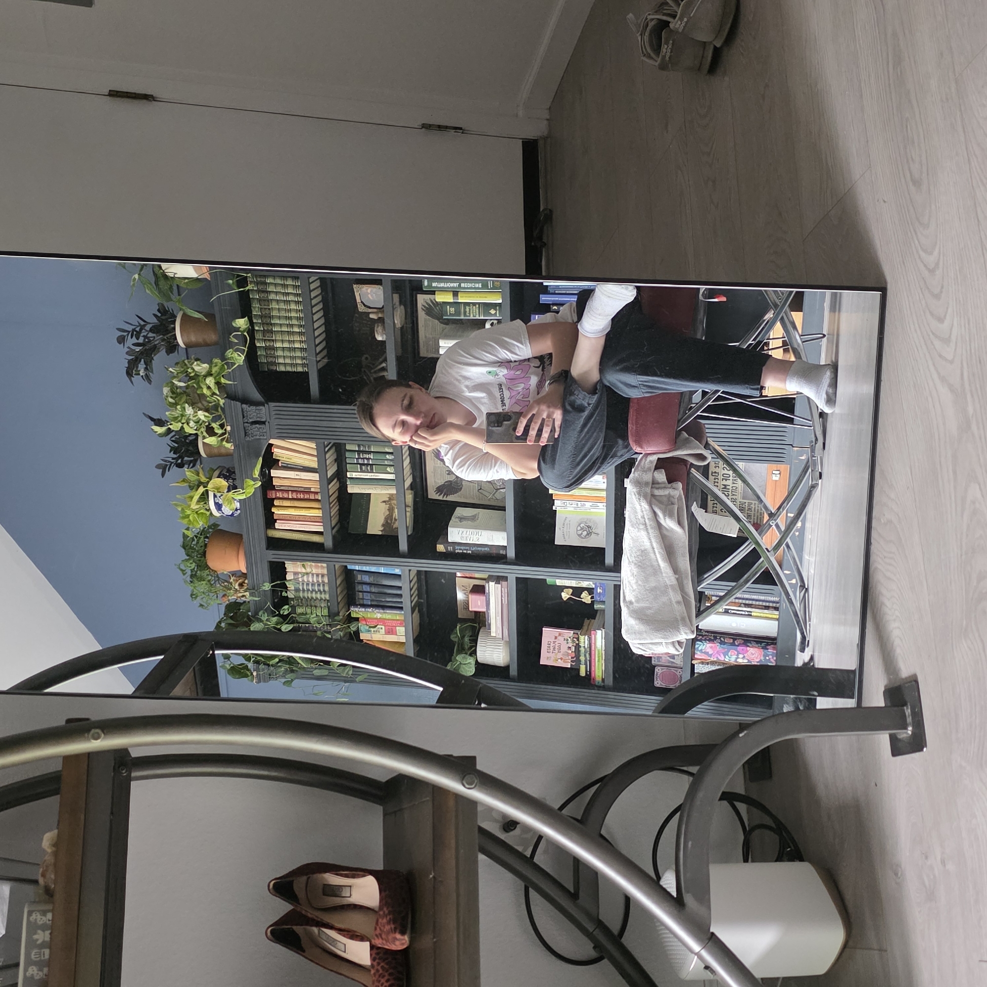 A person is seen in a full-length mirror, sitting on a red stool with a white towel draped over it. They are wearing a white t-shirt and dark pants, with white socks on their feet. The person is holding a smartphone and appears to be taking a selfie. The mirror reflects a bookshelf filled with books and plants. To the left of the mirror, there is a metal shelving unit with two pairs of high-heeled shoes on the top shelf. The floor is light-colored wood. The scene is likely indoors in a residential setting in Portland, United States of America.