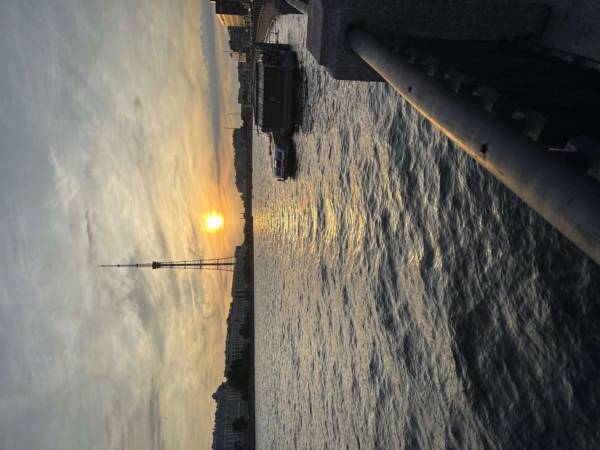 This is a scenic sunset view over the Neva River in Saint Petersburg, Russia. The sun is low on the horizon, casting a golden glow on the water and the sky, which is filled with wispy clouds. A tall television tower dominates the skyline in the distance, silhouetted against the bright sky. Several buildings, likely historic, line the riverbank, with a more modern structure visible on the right. Two boats are moored in the river, one of which appears to be a floating house or structure. A stone embankment with a metal railing occupies the foreground, suggesting a promenade or viewing area. The water is calm, with gentle ripples reflecting the sunset light. The overall atmosphere is serene and picturesque, capturing a tranquil moment in the city.