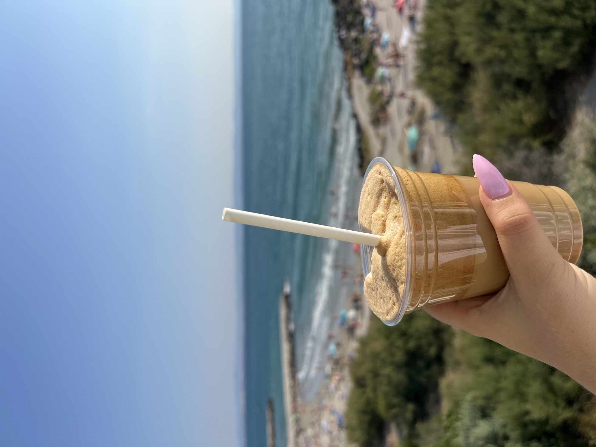 A hand with a light pink, almond-shaped fingernail holds a clear plastic cup filled with a frothy, light brown beverage and a white straw. The cup is positioned in the foreground, with a blurred background that depicts a sunny beach scene. The sea, a vibrant blue, stretches towards the horizon under a clear blue sky. A pier extends into the water, and the beach is populated with numerous people relaxing under umbrellas and sunbathing. Green bushes are visible in the mid-ground, framing the beach and sea. The overall impression is one of a refreshing drink enjoyed with a scenic view of the Black Sea coast in Pomorie, Bulgaria.