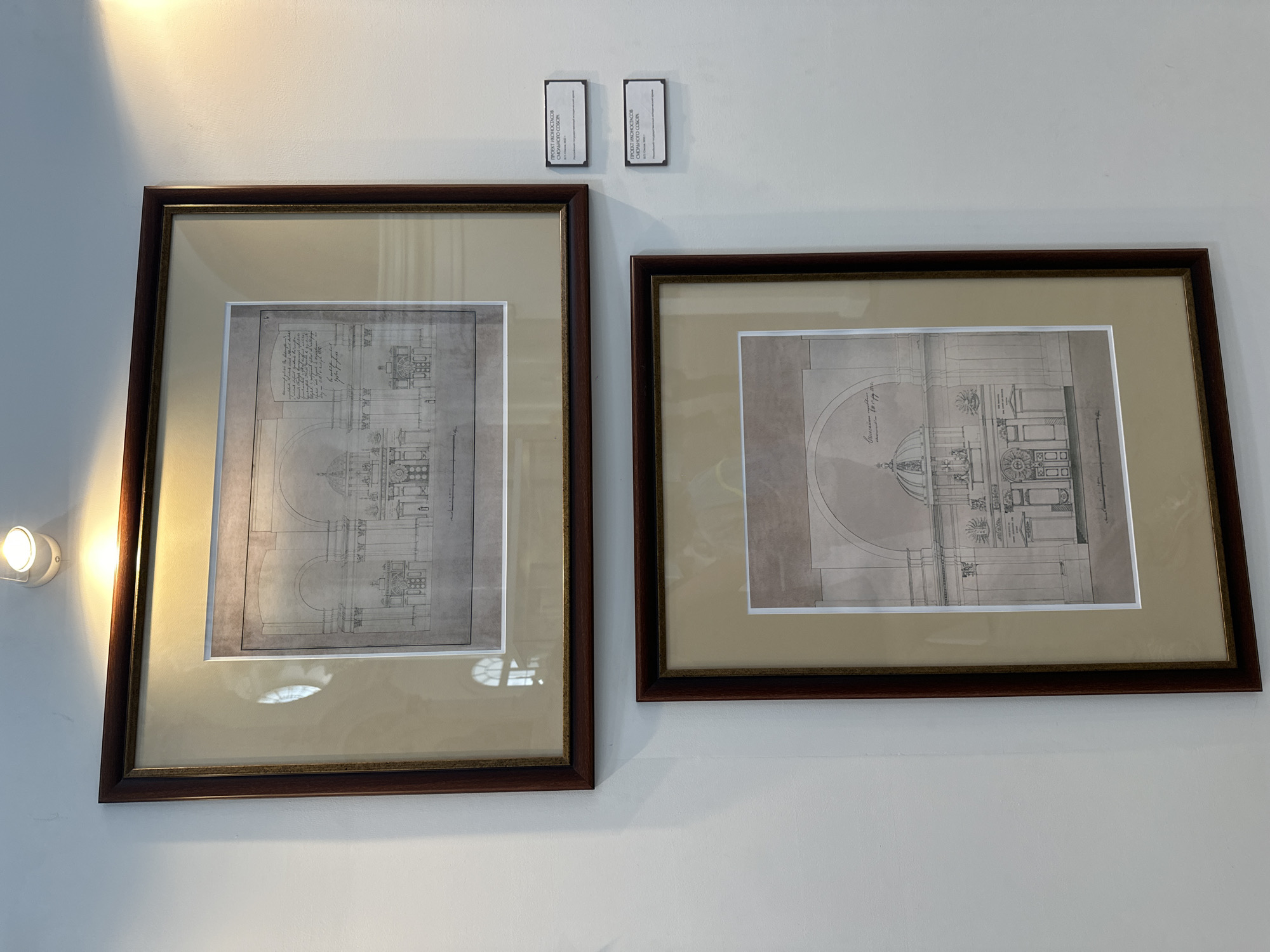 Two framed architectural drawings are displayed on a white wall in what appears to be a museum or gallery. The drawings depict elevations or facades of buildings, rendered in black and white with fine linework. Both drawings are matted with a light beige material and framed in dark wood. The upper drawing features a symmetrical building facade with multiple arches, columns, and ornate details, as well as some handwritten text. The lower drawing also shows a building facade, this one with a prominent dome and an arched entrance, also with handwritten annotations. Two small rectangular plaques with text are attached to the wall to the right of the framed drawings. The upper plaque is positioned to the right of the upper drawing, and the lower plaque is to the right of the lower drawing. The drawings and plaques are illuminated by spotlights from above, creating highlights on the wall and the framed artwork. The overall environment suggests an exhibition focused on historical architecture or design.