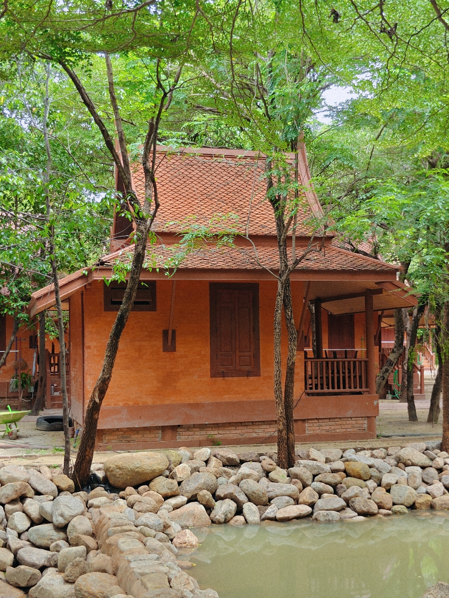 The image depicts a quaint, rustic building situated in a natural setting, likely a resort or traditional housing complex. The building itself is constructed with orange-brown brick walls and a characteristic tiered roof with orange tiles, suggesting Southeast Asian architecture. A wooden porch with a railing runs along the front of the building, and a single wooden door is visible. The building is surrounded by lush green trees with visible branches and leaves, indicating a tropical or subtropical environment. In the foreground, a body of murky green water is bordered by a collection of large and small stones, forming a naturalistic edge. The overall impression is one of a serene and tranquil location, possibly in a rural or semi-rural area.