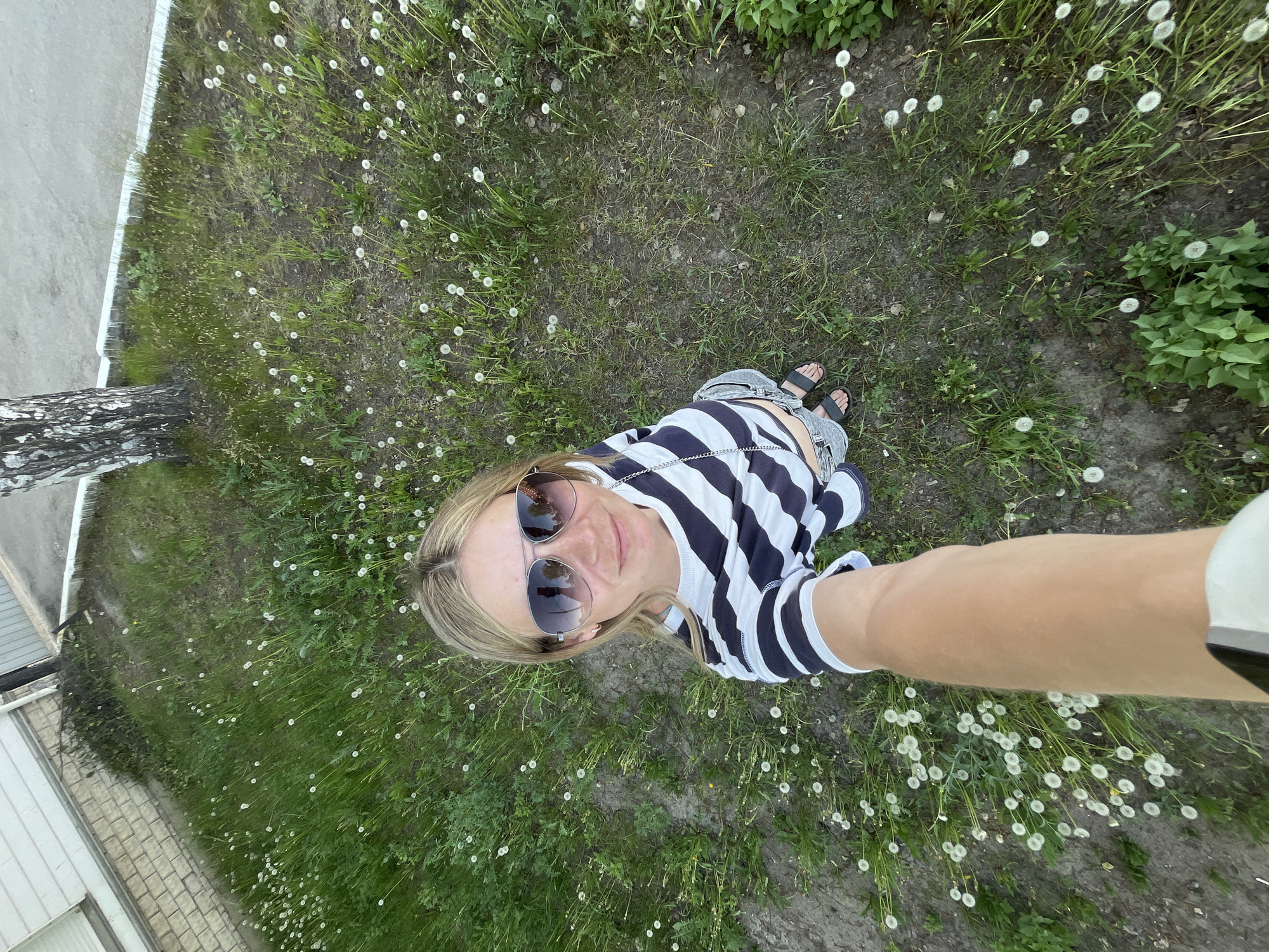 A woman stands on a grassy area with numerous dandelions, taking a selfie. She is wearing a striped shirt, jeans, and sandals. Sunglasses are perched on her head. Part of a building and a tree are visible in the periphery. The location appears to be an outdoor area in the city of Chaikovsky, Russia, based on the image's metadata.