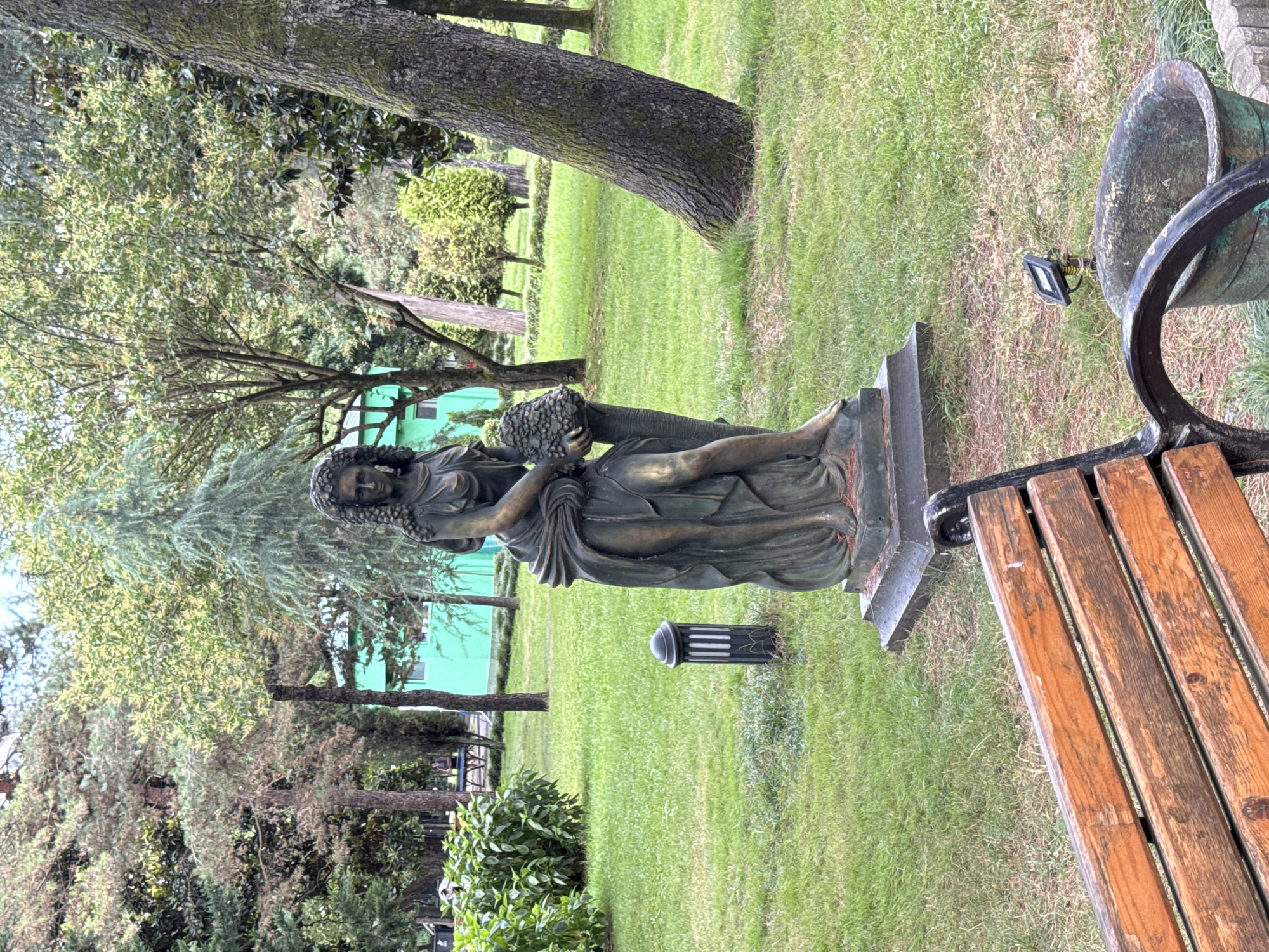 A bronze statue of a woman holding a bunch of grapes stands in a park in Batumi, Georgia. The statue is on a dark pedestal in a grassy area with trees and a green building in the background. A wooden bench is in the foreground, with a planter and a small solar light nearby. The day is overcast.