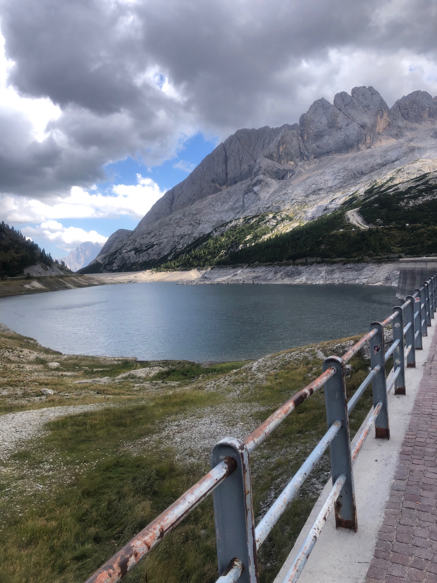 The image shows a panoramic view of the Fedaia Lake and the Marmolada mountain range, located in Canazei, Italy. In the foreground, a rusty metal railing runs along a paved walkway. The lake, with its calm, blue-green water, occupies the central part of the image, with the rugged, rocky slopes of the Marmolada mountain forming a dramatic backdrop. Dark clouds hover in the sky, casting shadows over the landscape. The slopes of the mountain are partially covered with sparse vegetation and trees. A concrete dam is visible on the right side of the lake, and a road with a few parked cars winds up the mountainside in the distance. The overall scene depicts a serene, yet majestic, natural landscape.