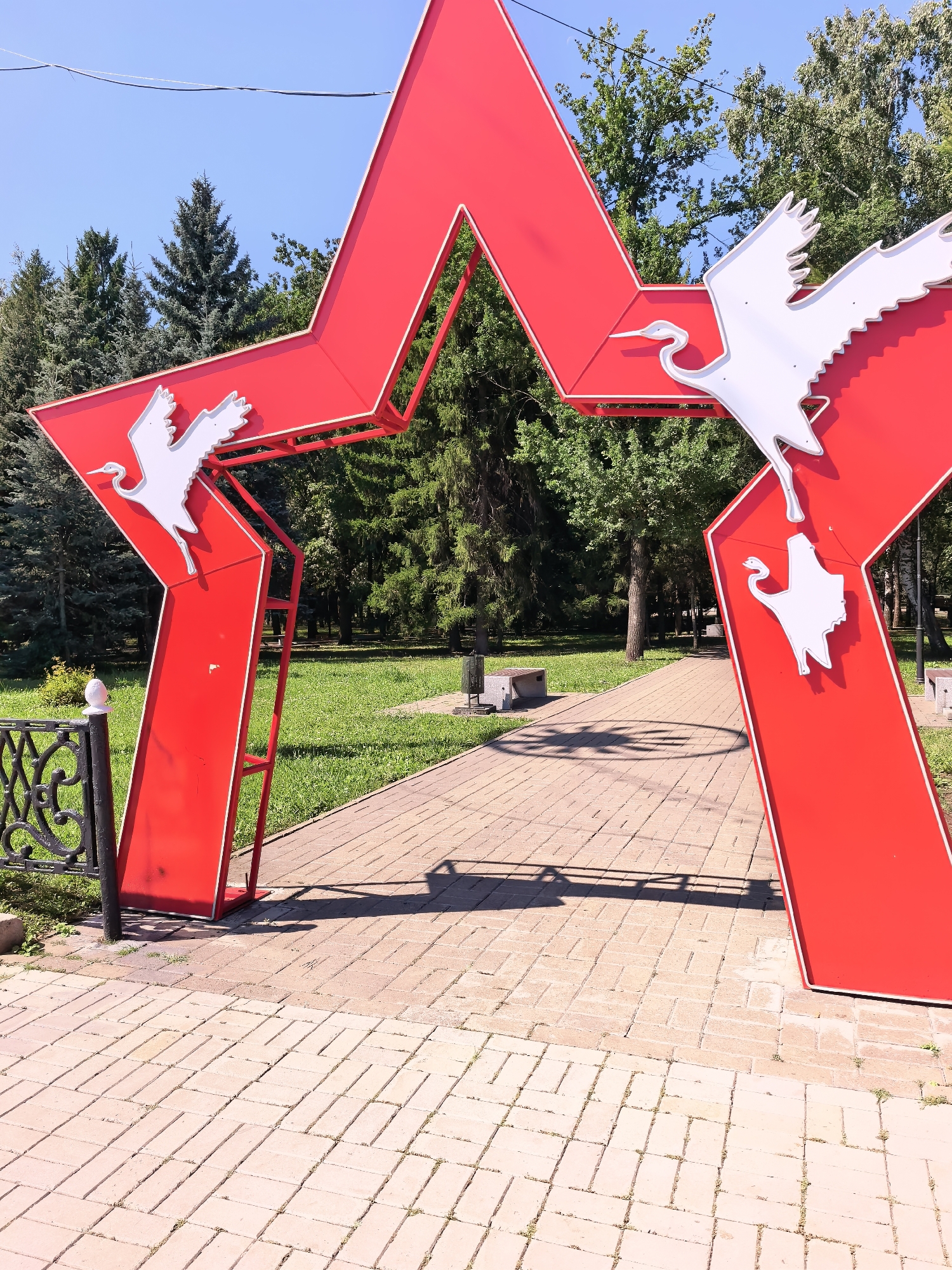 The image depicts a red star archway with white crane silhouettes adorning its points. The archway is situated in a park with a paved walkway leading through it. Trees and a clear blue sky form the background. This archway is located in the city of Sterlitamak, Russia, specifically in the memorial complex dedicated to the Great Patriotic War.