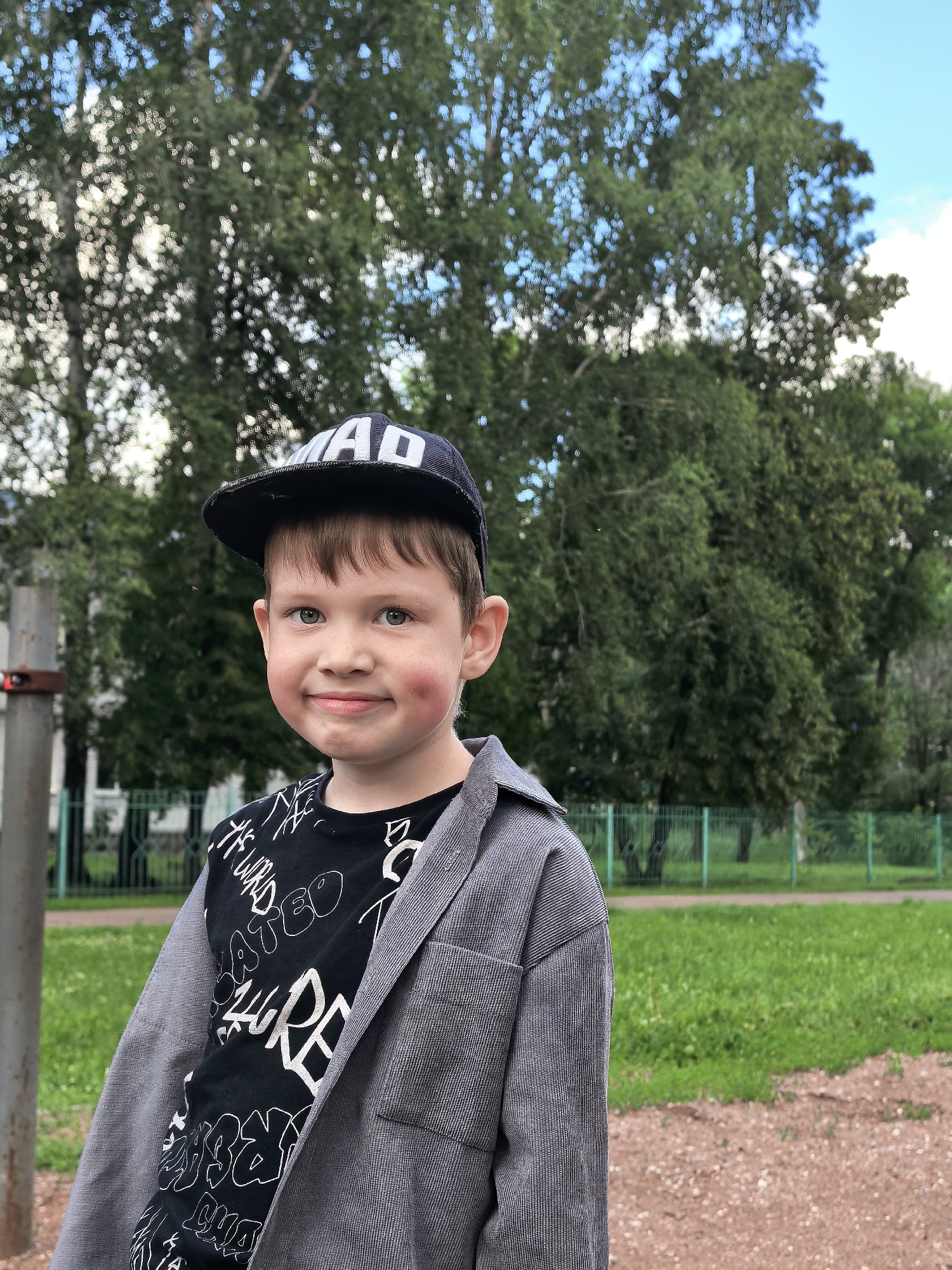 This is a medium shot of a young boy wearing a black baseball cap with white lettering that reads "MAD". He is also wearing a black t-shirt with white graffiti-style text and a gray, corduroy-like jacket. The boy has light brown hair and blue eyes, and he is smiling directly at the camera. His cheeks are rosy, and he has dimples.
The setting appears to be an outdoor park or playground. Behind the boy, there are green trees and a bright blue sky with some clouds. A green metal fence is visible in the background, and a small part of what looks like playground equipment is on the left side of the frame. The ground in the foreground is a mix of dirt and small stones.
The time of day appears to be daytime, likely afternoon, given the bright lighting. The weather seems pleasant, with clear skies. The overall mood of the image is happy and cheerful, conveyed by the boy's smile.
The visible text on the boy's cap reads "MAD". The text on his t-shirt is in a stylized font, appearing to be a mix of English words and possibly made-up words or artistic scribbles.