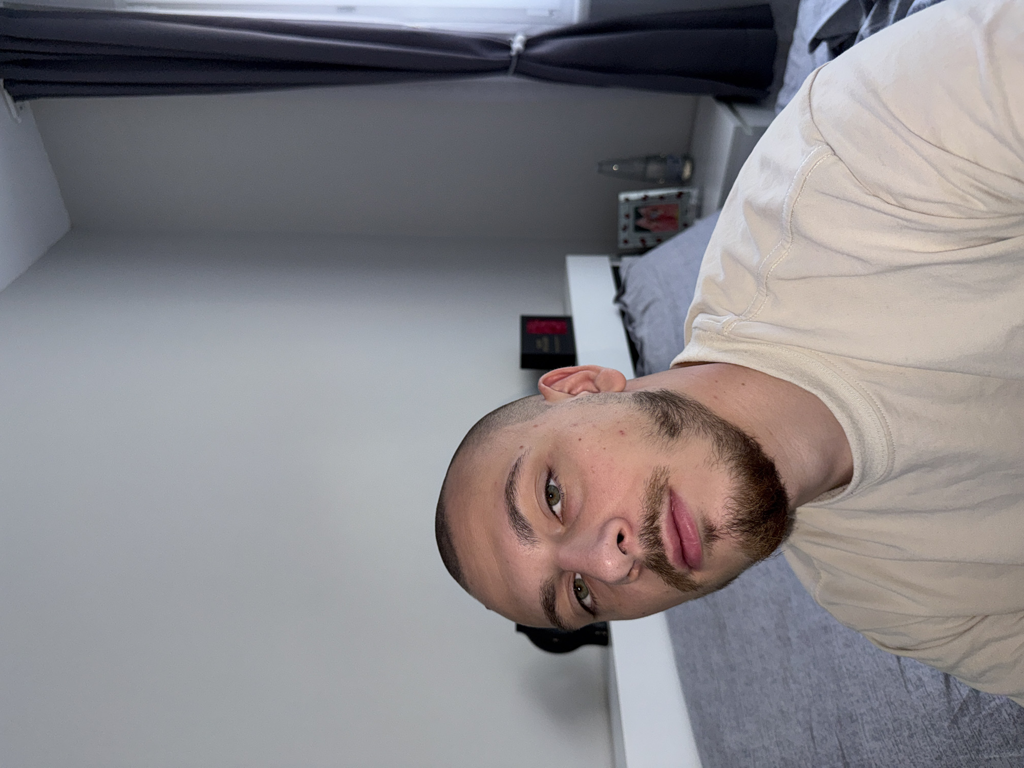 The image shows a young man with a shaved head and a goatee, wearing a light-colored t-shirt. He is looking directly at the camera with a neutral expression. The background is a bedroom with a grey duvet on a bed, a white headboard, and a bedside table with a framed picture and a bottle on it. A dark grey curtain is visible on the right side of the image, partially covering a window. The overall lighting suggests it is daytime, with light coming from the window. The scene is set indoors, in what appears to be a domestic environment. No specific landmarks or indicators place the location in Stara Zagora, Bulgaria; the description is based solely on the visual elements within the image.