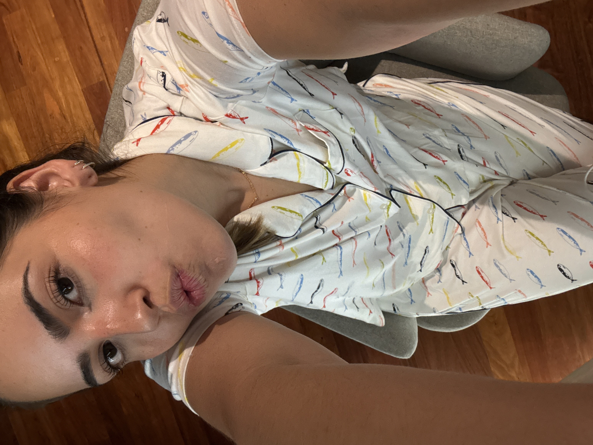 A woman is positioned at an angle, appearing to take a selfie. She is wearing white pajamas adorned with a pattern of colorful fish. Her lips are pursed in a kissing gesture. She is sitting on a gray chair. The background consists of a wooden floor. There is no indication of the location being in San Bernardino Tlaxcalancingo, Mexico.